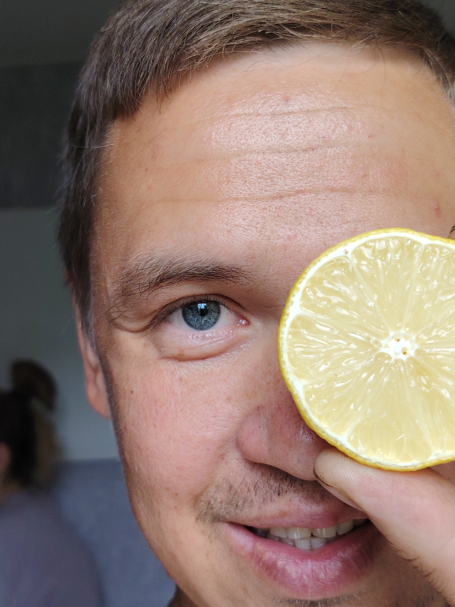 A close-up, low-angle, medium shot captures the right half of a light-skinned man's face. He has short, brown hair, visible stubble on his upper lip and chin, and a blue eye. A slice of lemon is positioned over his right eye, obscuring it from view. His mouth is slightly open, revealing his teeth. The background is blurred, showing a hint of a grey wall and what appears to be another person in the distance. The lighting is soft and natural. The image was taken in the city of Chaikovsky, Russia.