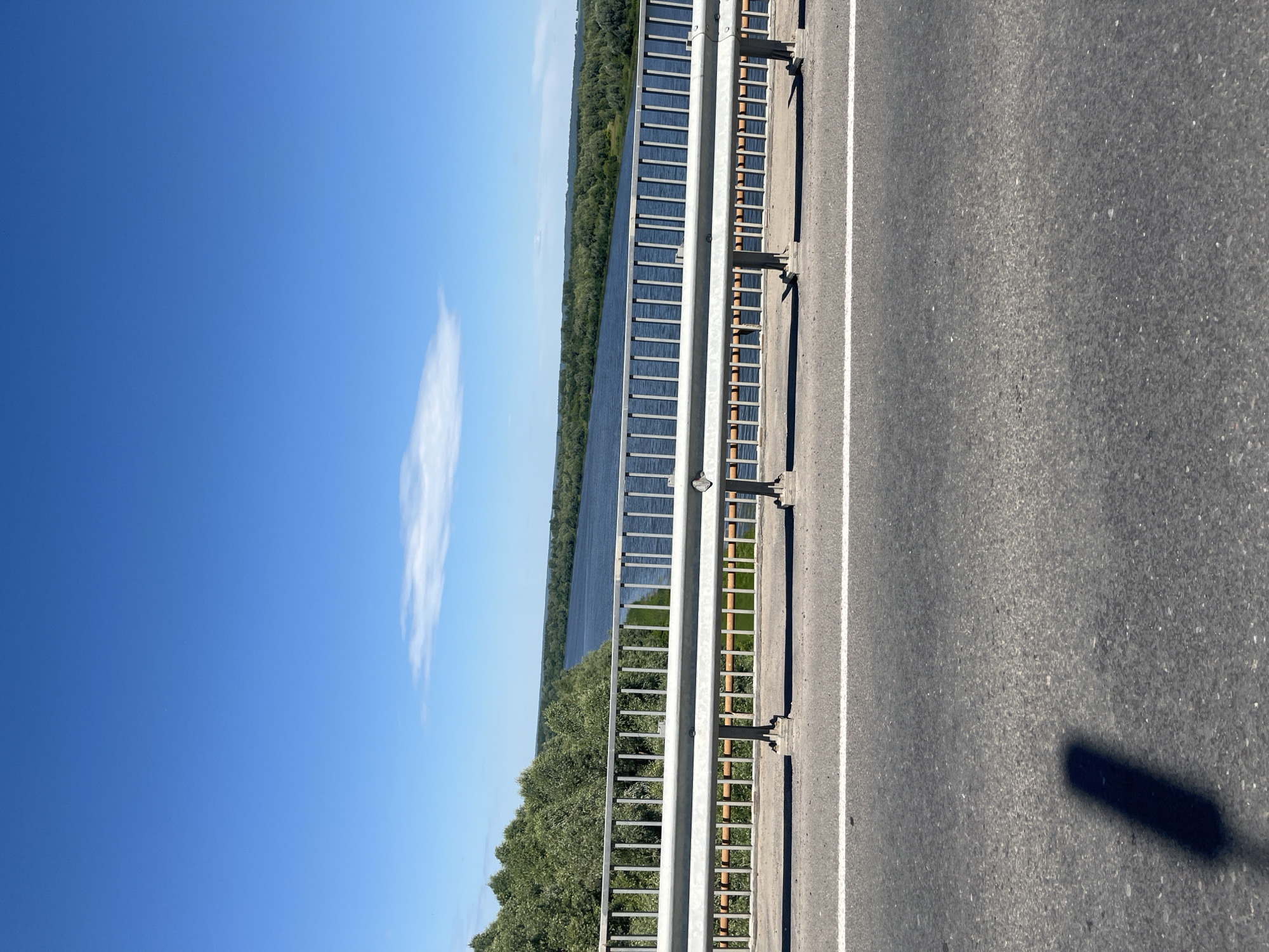 The image shows a view from a bridge over a wide river, likely the Volga, as seen from the city of Yadrin, Russia. The sky is a clear, vibrant blue with a few wispy clouds. A metal railing with vertical bars runs across the foreground, separating the road from the river view. The road surface is asphalt with a white line marking the lane. Lush green trees line the banks of the river, creating a dense, verdant landscape that stretches into the distance. The water is a deep blue and appears calm. The overall scene is one of natural beauty and tranquility.