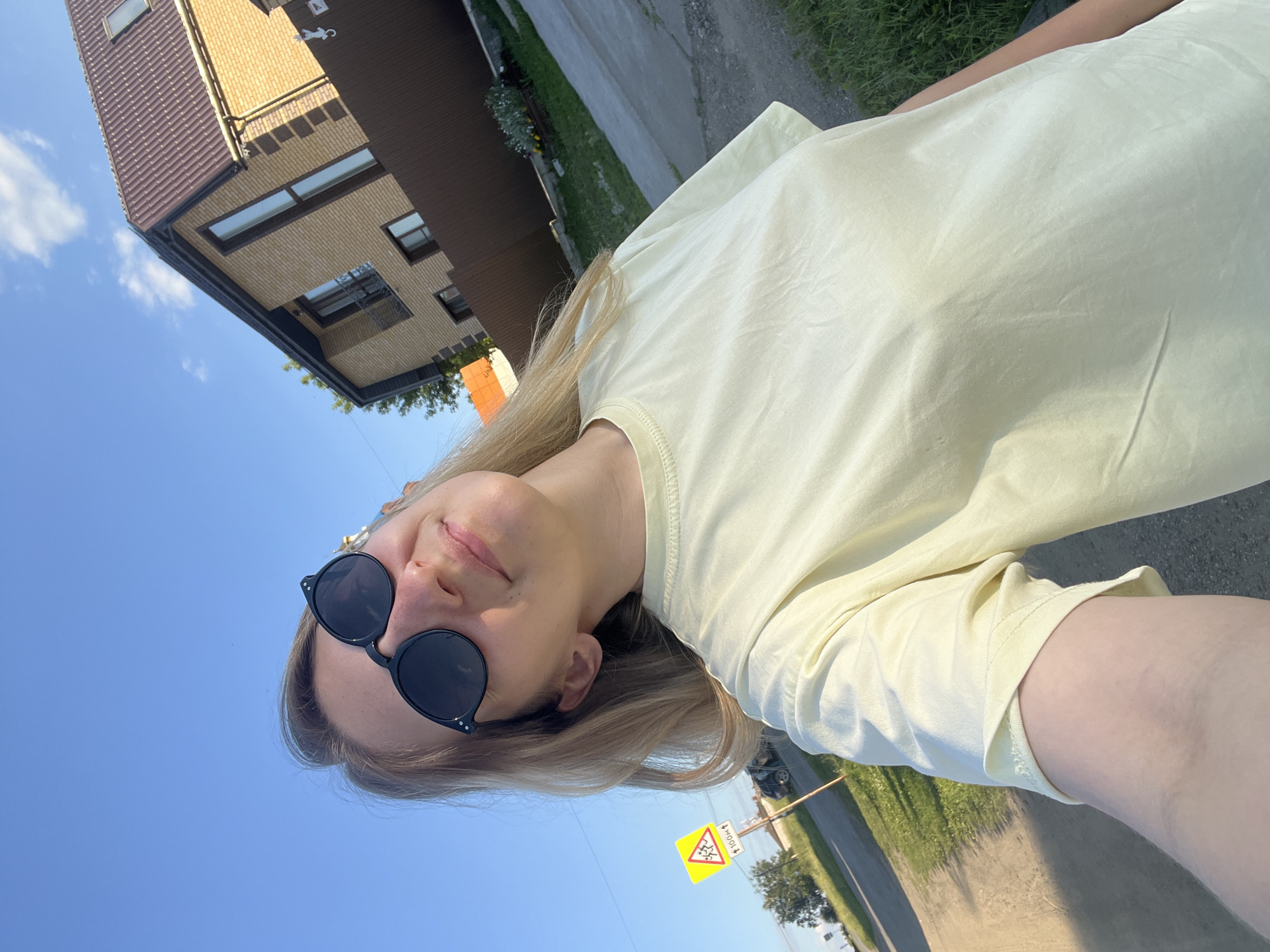 A young woman with blonde hair and sunglasses is taking a selfie outdoors. She is wearing a pale yellow t-shirt. In the background, there is a modern house with beige brickwork and a brown tiled roof. A paved driveway or road is visible, bordered by green grass and trees. The sky is clear and blue, suggesting it is daytime. A yellow triangular road sign with a black symbol is visible in the distance, indicating a hazard. The overall scene appears to be in a residential area in Бруснятское, Russia. The woman's expression is relaxed and happy.
