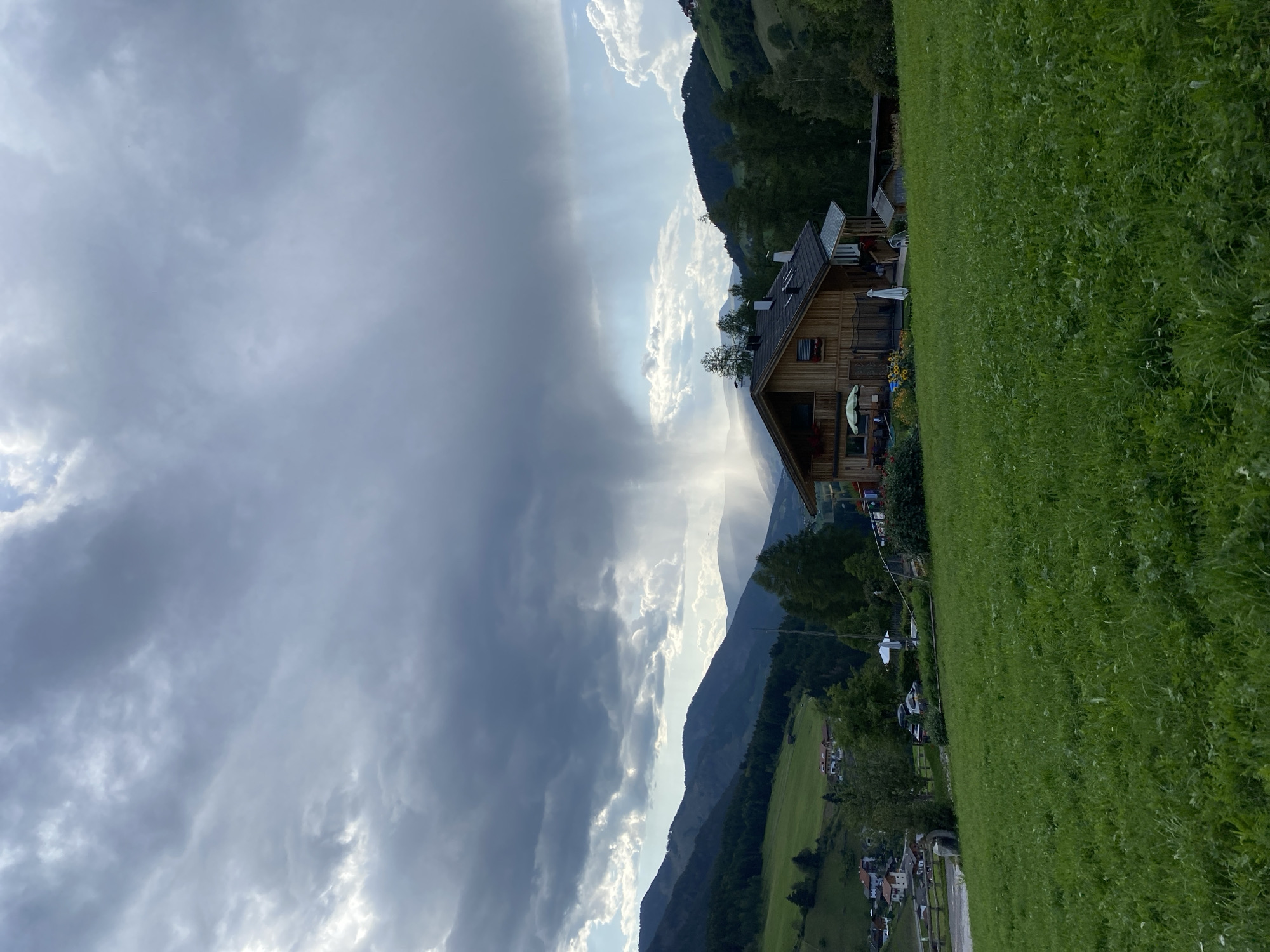 In the city of St. Magdalena - Santa Maddalena, Italy, a wooden house with a dark roof sits in the foreground, partially obscured by a grassy hill. The house has a balcony and several windows, and in front of it, a patio area is set up with outdoor seating and an umbrella. The house is surrounded by trees and flowerbeds. In the background, rolling hills and mountains are visible, with a dramatic sky above. A rain shower is occurring in the distance, with visible streaks of rain falling from dark clouds. The overall scene is one of a rural landscape with a rustic dwelling.