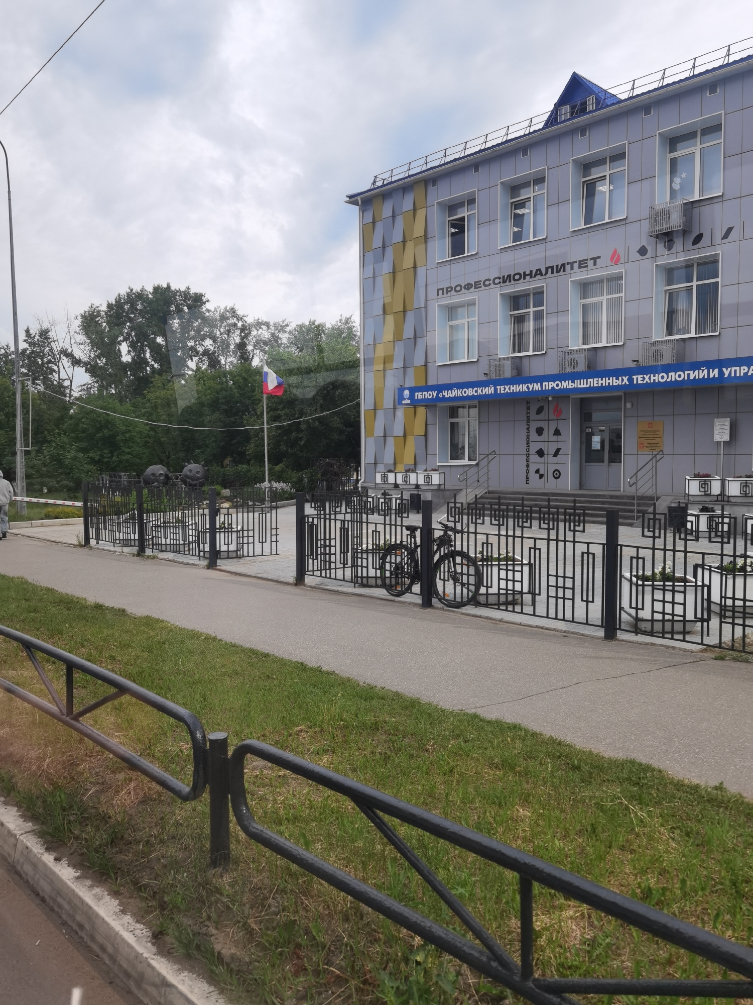 The image displays an exterior view of the State Budgetary Professional Educational Institution "Chaikovsky College of Industrial Technologies and Management" (ГБПОУ «ЧАЙКОВСКИЙ ТЕХНИКУМ ПРОМЫШЛЕННЫХ ТЕХНОЛОГИЙ И УПРАВЛЕНИЯ») in Chaikovsky, Russia. The multi-story building features a light blue-grey facade, with a distinctive yellow and grey geometric pattern covering part of its left side. A prominent sign on the building reads "ПРОФЕССИОНАЛИТЕТ" (PROFESSIONALITET), accompanied by a flame-like logo. Another vertical sign next to the main entrance also bears the "ПРОФЕССИОНАЛИТЕТ" text, surrounded by various industrial-themed geometric icons.
In front of the college, a paved area leads to the main entrance. This area is bordered by an ornate black metal fence, against which a black bicycle is parked. White planters containing greenery are arranged along the fence. To the left of the entrance, a Russian flag (white, blue, red horizontal stripes) flies on a flagpole, with two dark, spherical, abstract sculptures visible nearby.
In the foreground, a simpler black metal barrier separates a strip of green grass from a paved path. A person wearing a light-colored hoodie is partially visible walking on the far left. The scene is set during the daytime under an overcast sky, with abundant green trees forming a backdrop. A tall black street light stands on the far left. No specific activities or events are prominently taking place, suggesting a quiet, typical day at the institution.