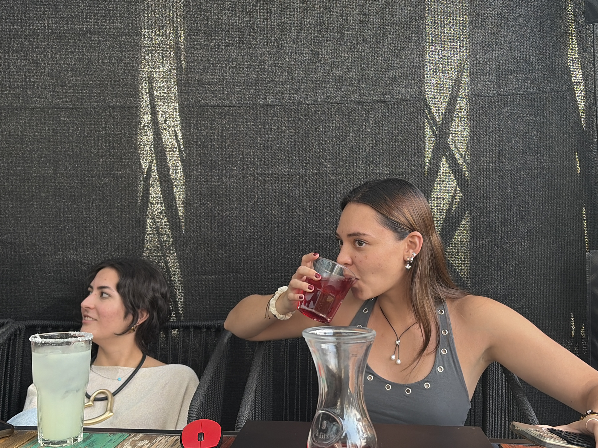 The image depicts two women seated at a table in an outdoor setting, possibly a restaurant or cafe. The woman on the right is drinking from a clear glass containing a red liquid. She has long brown hair, fair skin, and is wearing a grey tank top. She also has jewelry including earrings and a necklace. The woman on the left has short dark hair and is wearing a light-colored shirt. She has a beverage in a glass with a salted rim, and the table also features a clear pitcher, a red computer mouse, and a mobile phone. The background is a dark, textured wall, possibly made of woven material. The location is the city of Unknown, Unknown.