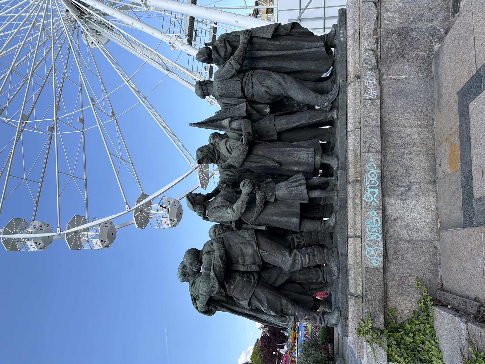 The image shows the Monument of Soviet Army in Sofia, Bulgaria, from a low-angle view with a Ferris wheel visible in the background. The bronze figures on the monument depict people in what appears to be civilian and military attire. The monument's base is made of stone with some graffiti present on the side facing the viewer. The sky is clear and blue. There is some greenery visible near the base of the monument. In the very background is what appears to be a fairground.