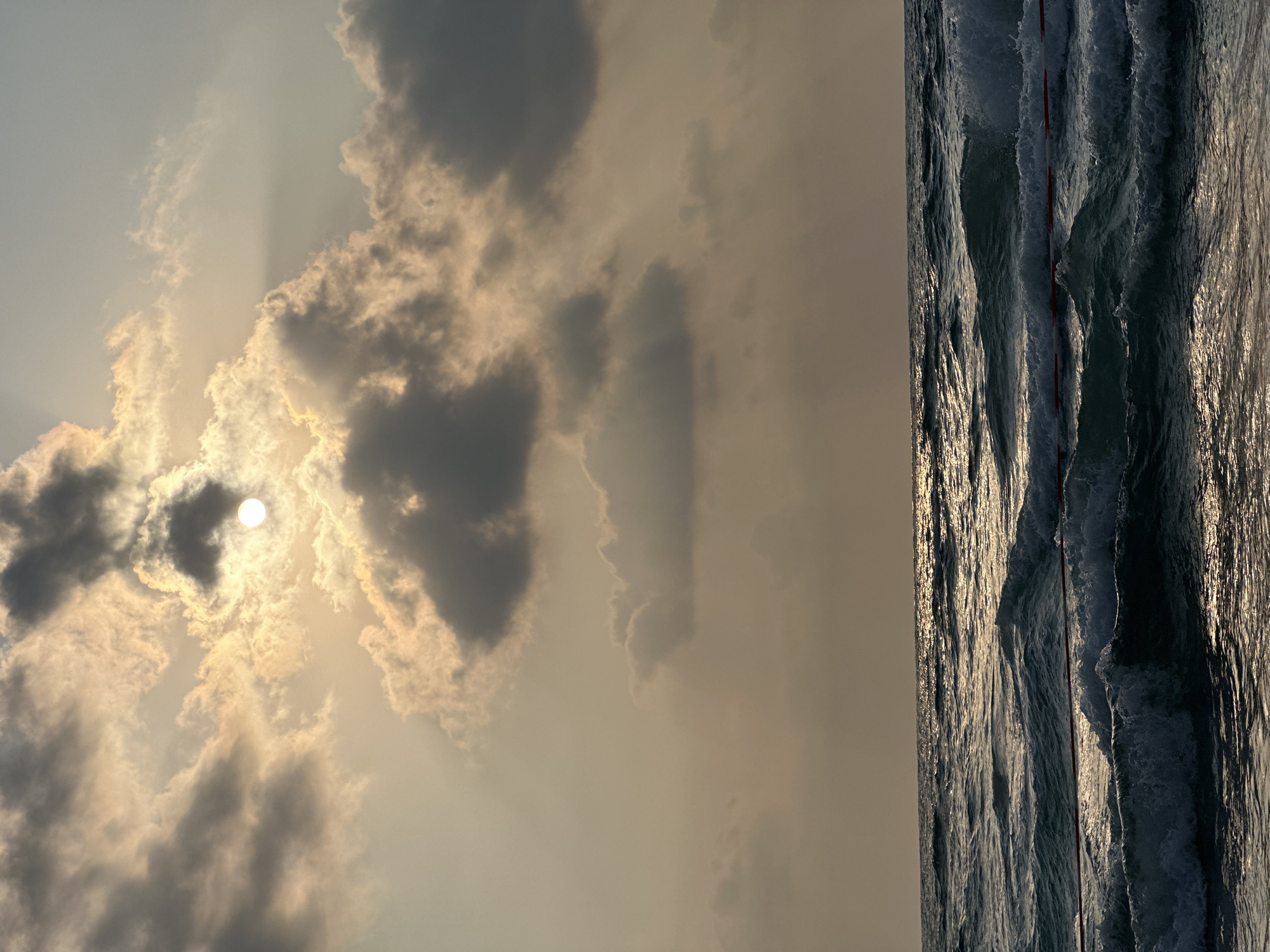 The image depicts a turbulent sea at Bat Yam, Israel, under a sky filled with dramatic clouds. The sun is partially obscured by these clouds, casting rays of light that create a shimmering effect on the water's surface. The waves are rough, with white caps indicating strong currents. A red line, possibly a marker or boundary, is visible in the foreground, partially submerged by the waves. The overall scene suggests a dynamic and somewhat wild coastal environment.