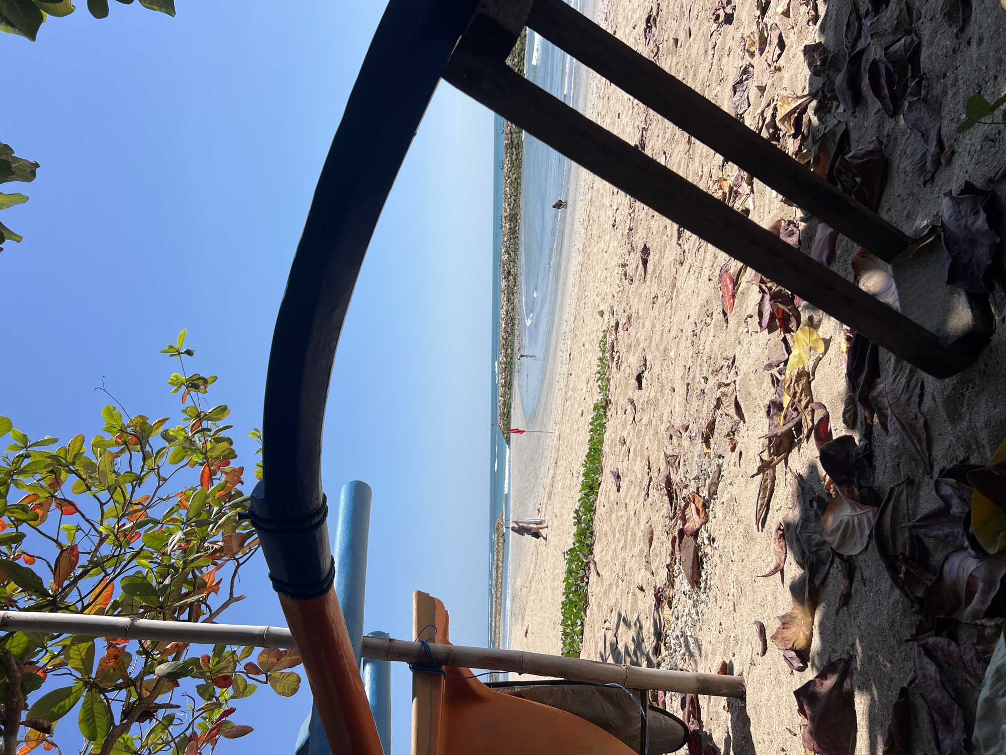This image depicts a sunny beach scene in Kuta, Indonesia, viewed from under the shade of a tree. In the foreground, fallen leaves and debris litter the sandy ground. Partially visible in the lower left is the orange hull of a boat, supported by a bamboo pole. A curved dark object, possibly part of the boat or a nearby structure, arcs across the upper portion of the frame. Beyond the immediate foreground, the beach stretches out, with a few people visible in the distance. The sea, with gentle waves, meets the shore, and a breakwater made of rocks extends into the water. Several small boats are dotted on the horizon. The sky is a clear, vibrant blue.