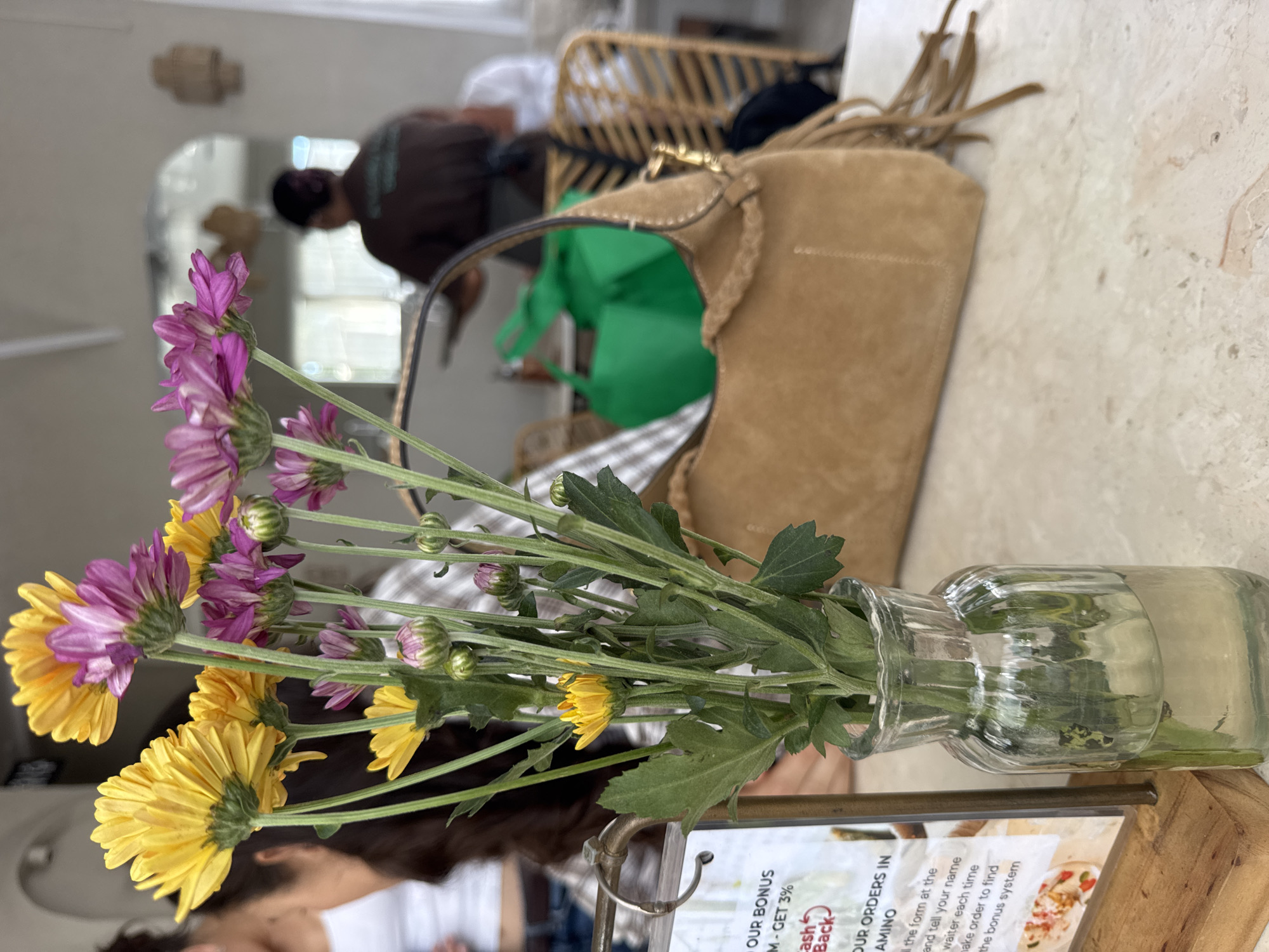 The scene captures an interior setting, likely a cafe or restaurant, as evidenced by the presence of a table, flowers in a glass vase, a menu card, and background elements suggesting a dining area. In the foreground, the vase contains a bouquet of yellow and purple flowers with green stems and leaves. Adjacent to the vase is a menu card displaying text, including "OUR BONUS," "GET 3%," and "OUR ORDERS IN."
In the middle ground, a brown suede handbag rests on the table, adding a touch of personal style. Behind the handbag, a green bag is visible, adding a pop of color. In the background, reflections in a mirror reveal a figure of a person wearing a brown shirt and a hat. A woven chair with tassels is also present. The overall environment appears to be a casual and stylish dining space. The location, Ungasan, Indonesia, is given.