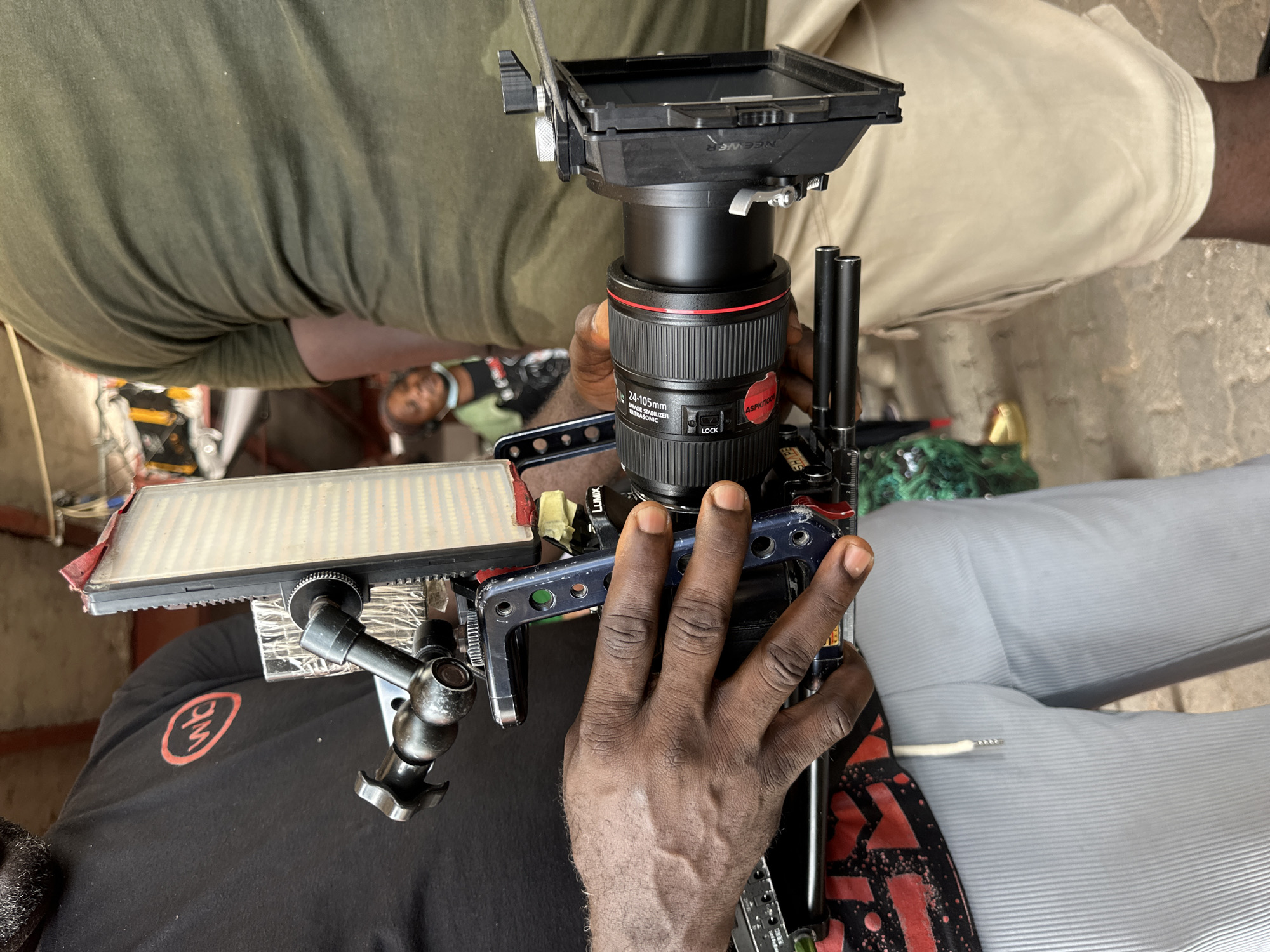 A close-up shot depicts a person of African descent holding a professional camera rig. The camera is equipped with a lens featuring a red ring, a matte box, and various mounting hardware. The rig includes a small LED panel light attached via an articulated arm. The individual's hand is prominently displayed holding onto the camera's support cage. The person wearing the black shirt has a red emblem on the front. The background includes portions of other people and clothing. The overall setting implies a film or video production environment. There is no explicit indication of the location being in Lagos, Nigeria, but this could be inferred from the subjects' ethnicity.