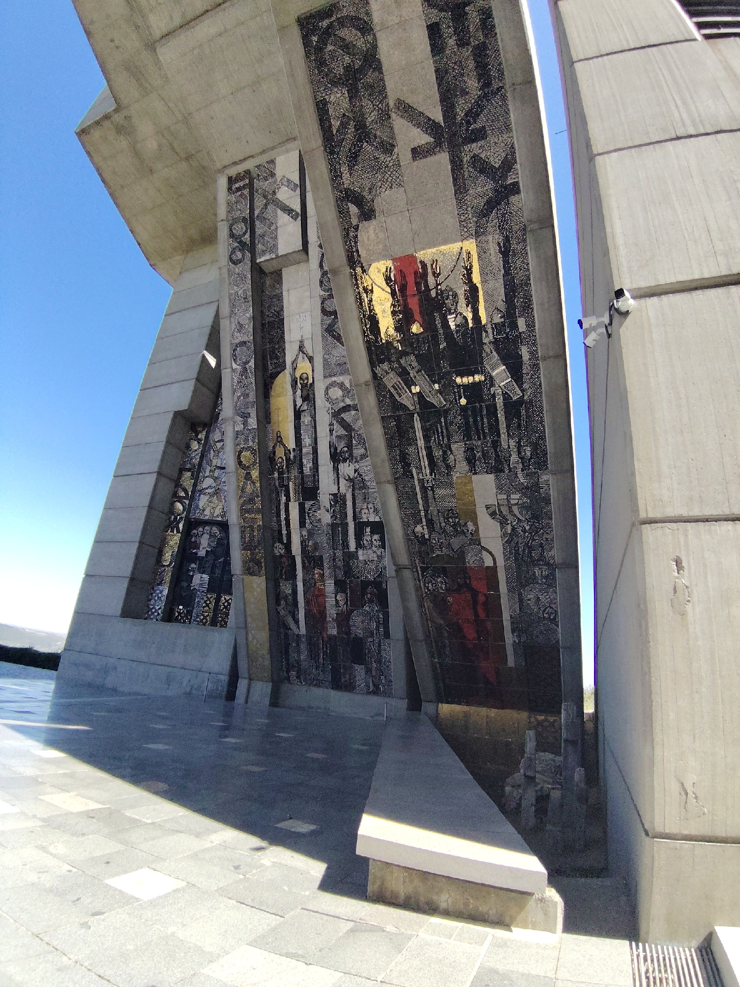 The image captures a low-angle perspective of the Founders of the Bulgarian State Monument, located in Shumen, Bulgaria. The monument features prominent concrete structures and decorative mosaic panels depicting historical figures and events. The panels are adorned with a variety of colors, including gold, red, black, and white, which adds to the monument's imposing aesthetic. The sky is visible as a gradient of blue, which contrasts sharply with the gray concrete and the complex mosaic designs. A surveillance camera is attached to one of the concrete pillars. The foreground shows a paved area, with a step leading towards the monument's base. The monument's scale is emphasized by the perspective.