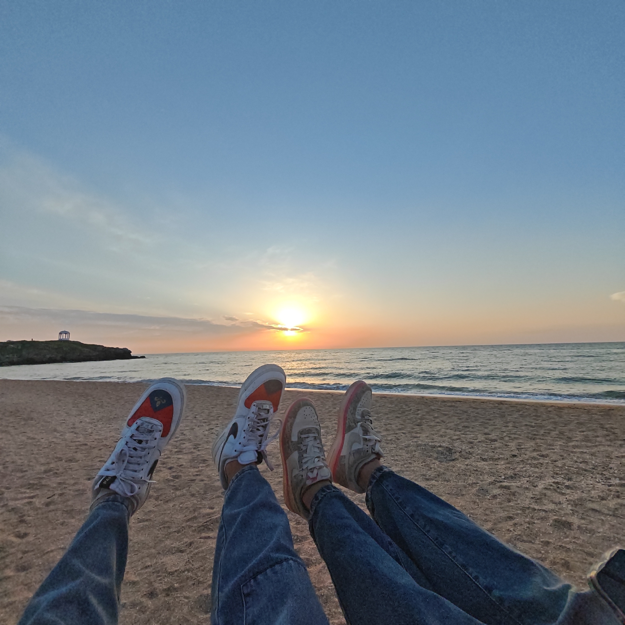 Two people are sitting on a beach at sunset. They are wearing jeans and sneakers. Their legs are extended in front of them. The sea is in the background, and the sun is setting over the horizon. A gazebo-like structure is on a rocky outcropping in the distance on the left side of the image. The scene appears to be a beach in городское поселение Щелкино, Ukraine, based on the location.