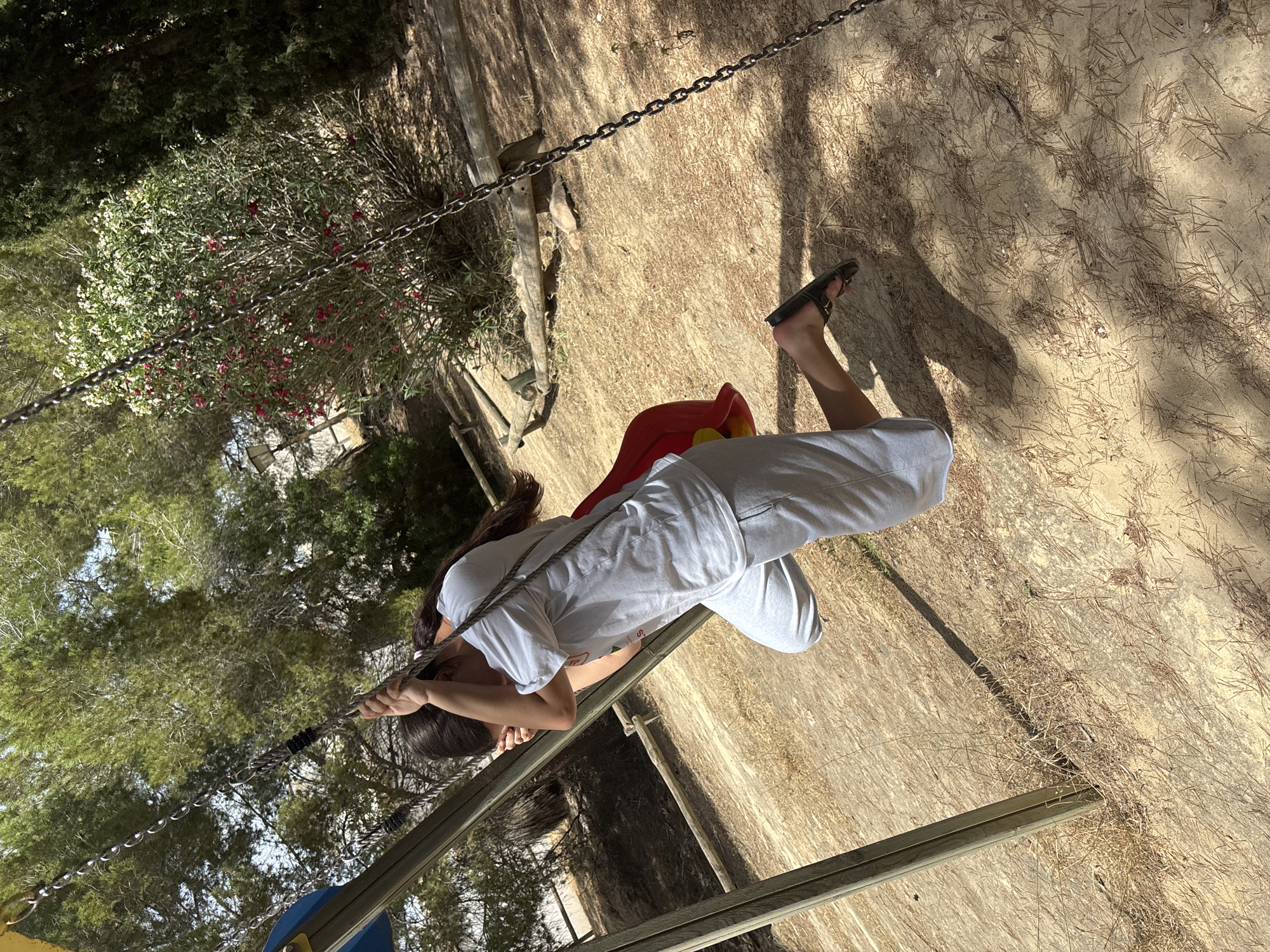 A young woman is pictured on a swing set, her body angled towards the ground. She wears a white t-shirt and light gray sweatpants, with dark hair falling over her shoulders. Her hands grip the swing chains, and she is wearing black flip-flops. The swing seat appears to be red and yellow, suggesting it's part of a playground. The background consists of dry, sandy ground with scattered pine needles and patches of green foliage and trees. Sunlight filters through the trees, casting dappled shadows on the ground, indicating a sunny day. The overall setting suggests an outdoor recreational area, possibly a park or playground, in a warm climate, consistent with the location context of Orihuela, Spain. There is no visible text in the image.