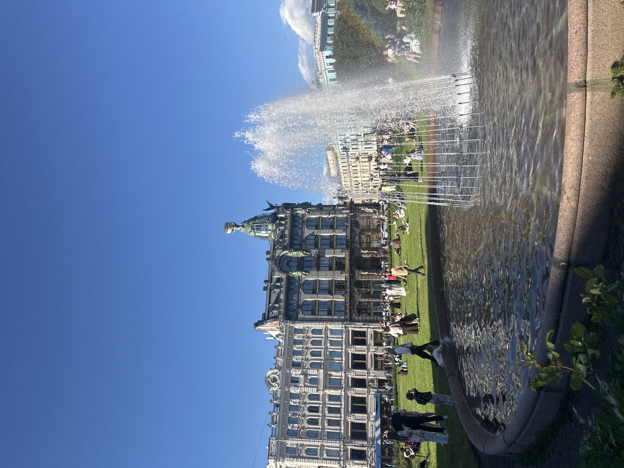 In Saint Petersburg, Russia, a wide shot captures a bustling city square on a clear, sunny day. A large fountain sprays water high into the air in the foreground, its water falling into a circular basin. Beyond the fountain, a green lawn is dotted with numerous people, many of whom are seated or picnicking. Several people are walking across the grass. Behind the lawn, grand, ornate buildings with multiple stories and arched windows line the square. One building in particular, with a distinctive green dome topped by a globe, stands out. The sky above is a vibrant, cloudless blue, with a faint rainbow visible near the fountain.