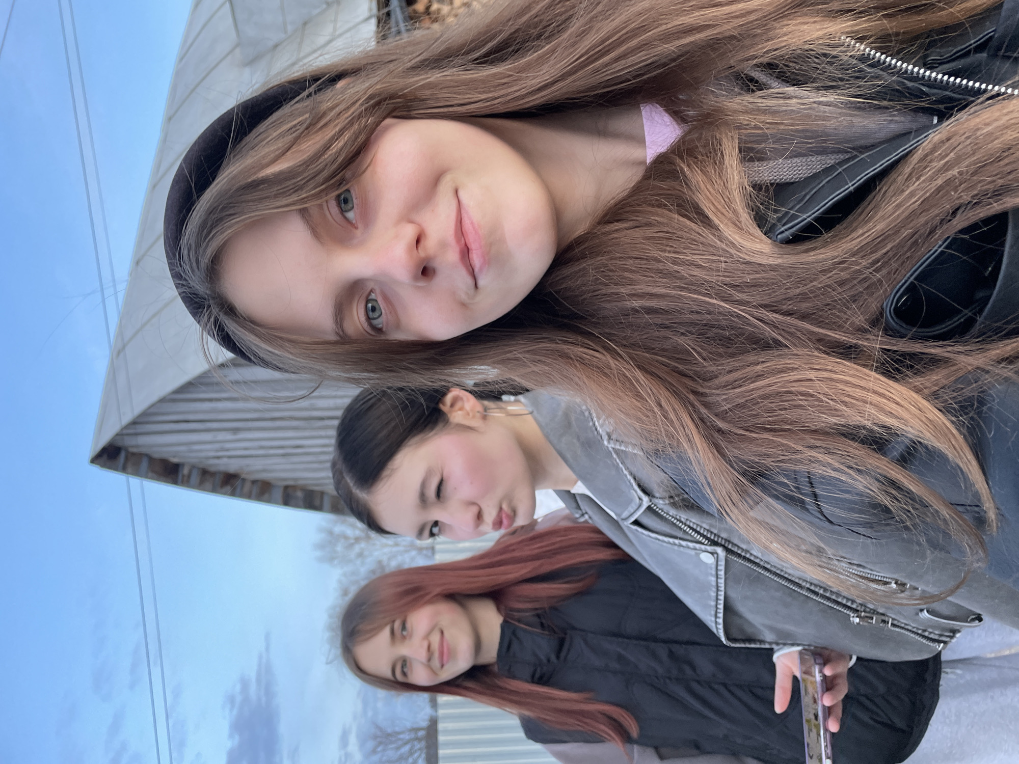 Three young women are posing for a selfie outdoors in Фоки, Russia. The woman at the top of the frame has long brown hair, blue eyes, and is wearing a black headband and a black jacket. The woman in the middle of the frame has dark hair pulled back, a grey jacket, and is winking and puckering her lips. The woman at the bottom of the frame has red hair and is wearing a black puffer jacket and is smiling. The background includes a cloudy sky, trees without leaves, and a corrugated metal fence. A building with a peaked roof is partially visible.