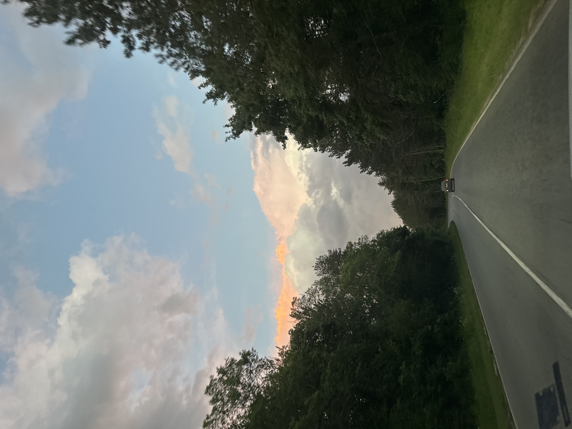 A slightly tilted, low-angle shot shows a road stretching away into the distance, framed by trees on either side. A car with illuminated tail lights is visible on the road. The sky above is partially cloudy, with hues of pale blue and pink suggesting a sunset or dawn. No people are visible in the image. The location is identified as Pryaporets, Bulgaria.