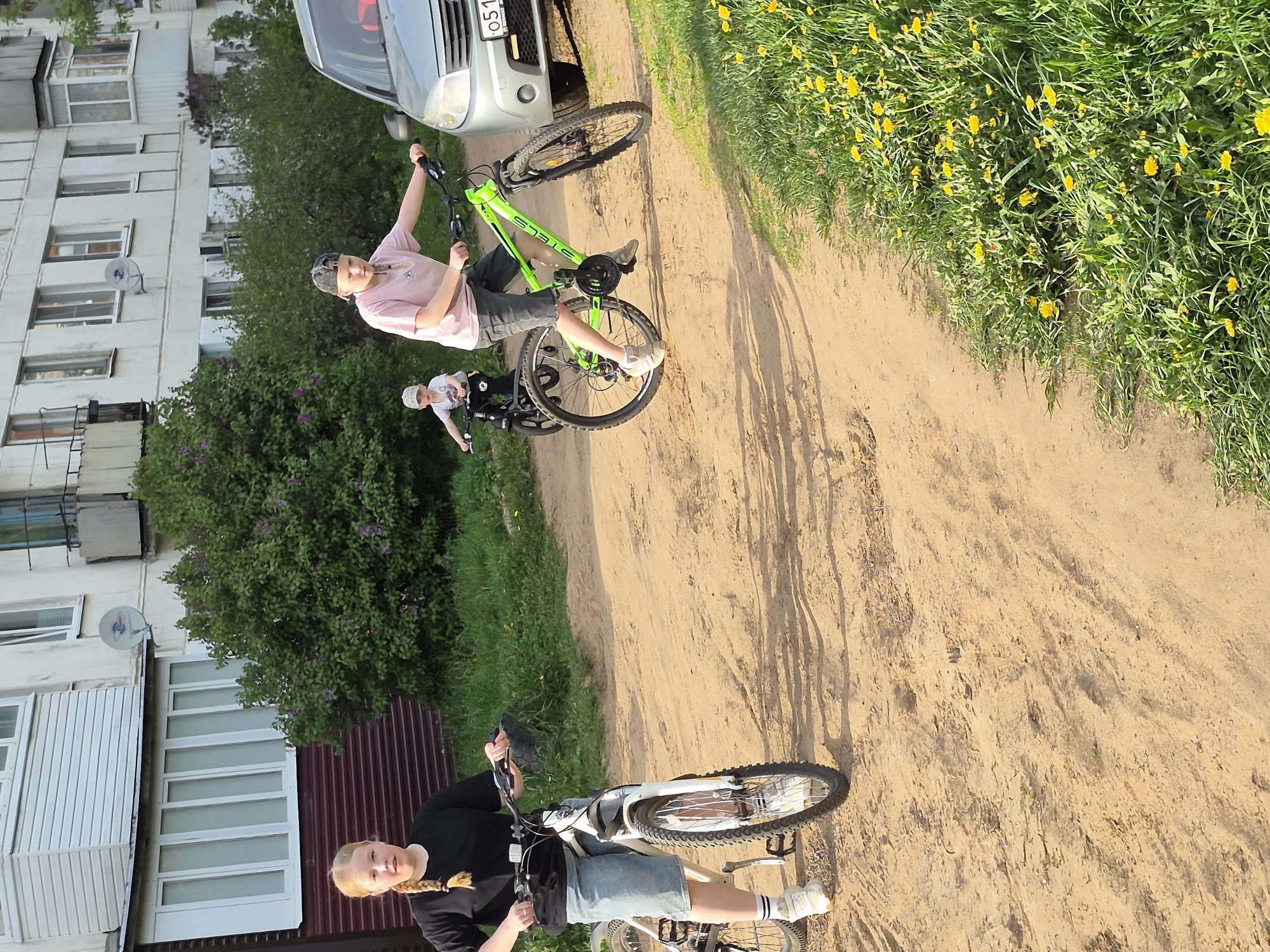 The scene captures three individuals on bicycles along a dirt path situated in an urban environment. The composition is characterized by a tilted perspective.
A fair-haired person, possibly female, is seen at the bottom of the frame, wearing a black long-sleeved shirt and denim shorts, mounted on a white bicycle. Above them, there is an individual in a pink shirt and dark shorts riding a green bicycle. Further up is another person wearing a white top and helmet, also on a bicycle.
In the background, there is a residential building. A parked car is situated beside the individual on the green bicycle. Yellow wildflowers and green foliage line the side of the dirt path.
