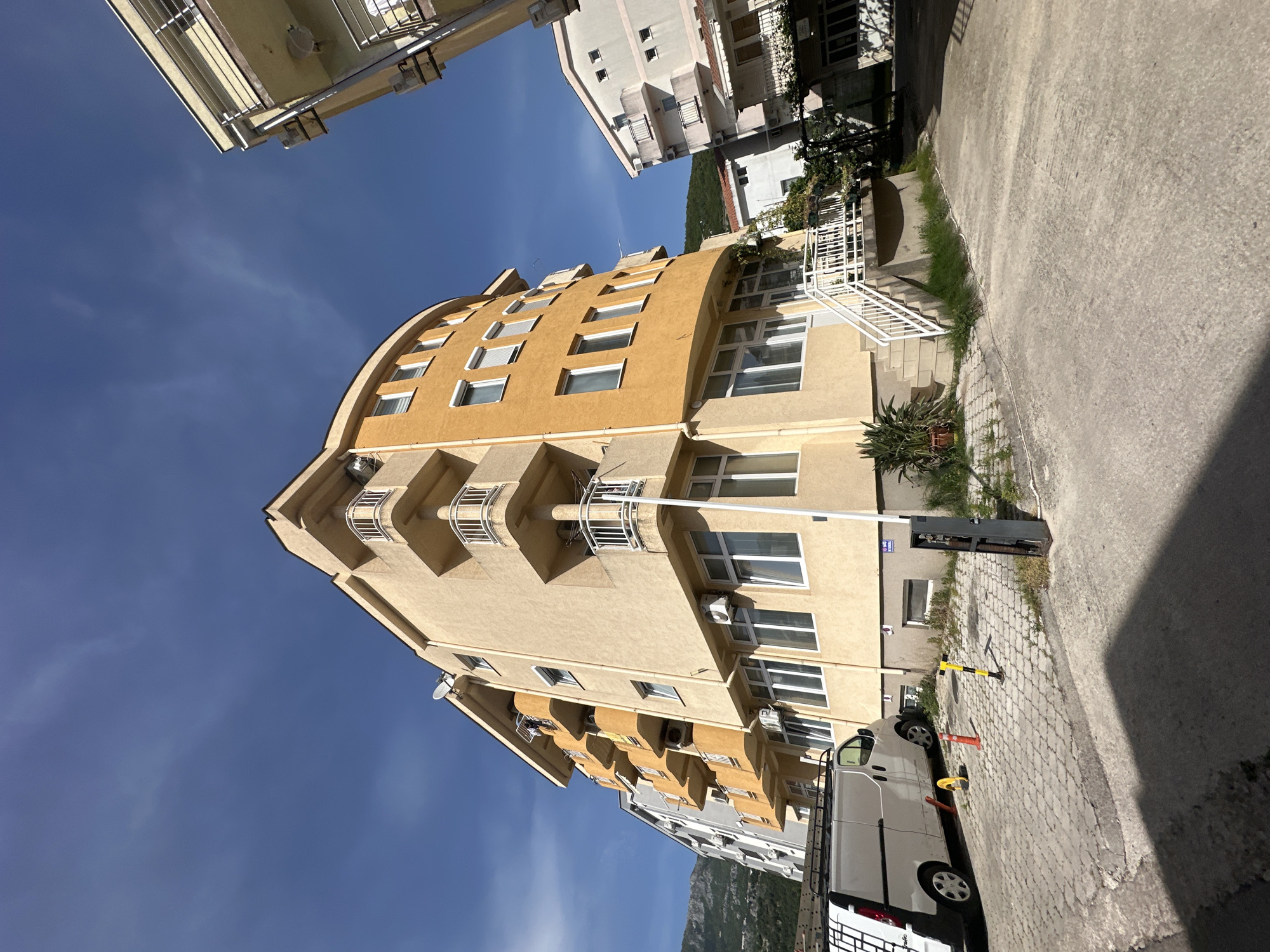 The image depicts a street scene in Budva, Montenegro, showcasing modern architecture against a backdrop of a clear, blue sky. An apartment building is featured prominently, its facade displaying a combination of light and dark yellow hues, adorned with symmetrical windows and balconies. Adjacent to the building, a paved street is visible, suggesting urban infrastructure. A white van is parked along the street, adding a sense of everyday activity to the scene. The overall composition presents a blend of residential and urban elements, common to this coastal city. The natural landscape is visible.