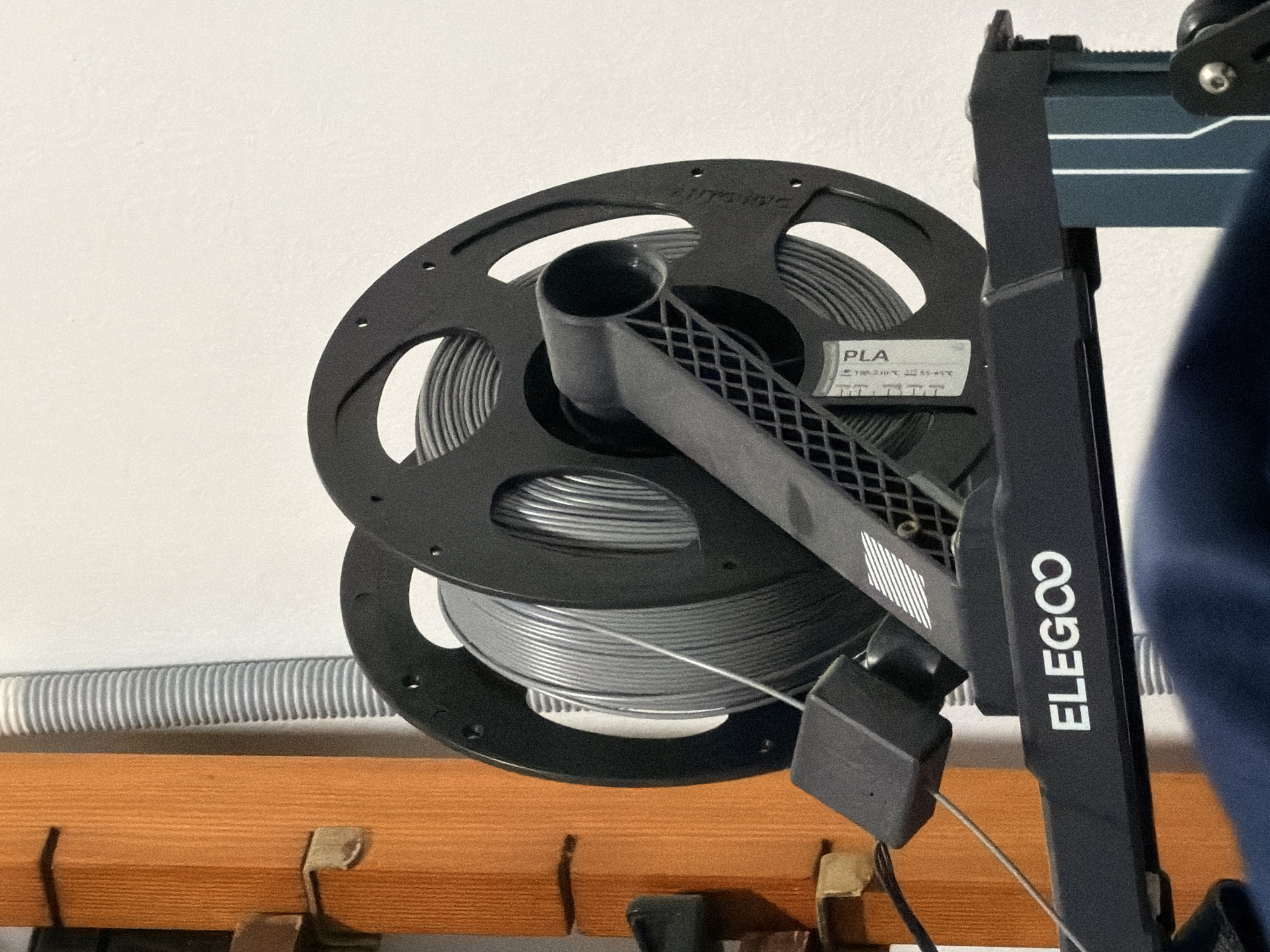 The image shows a spool of grey filament on a black 3D printer spool holder. The spool holder is branded with the ELEGOO logo. A grey corrugated tube is visible in the background, as is a wooden structure. The scene appears to be indoors, possibly in a workshop or home setting. There is no indication of the city of Torremolinos, Spain, or any specific location. No people or actions are depicted.
