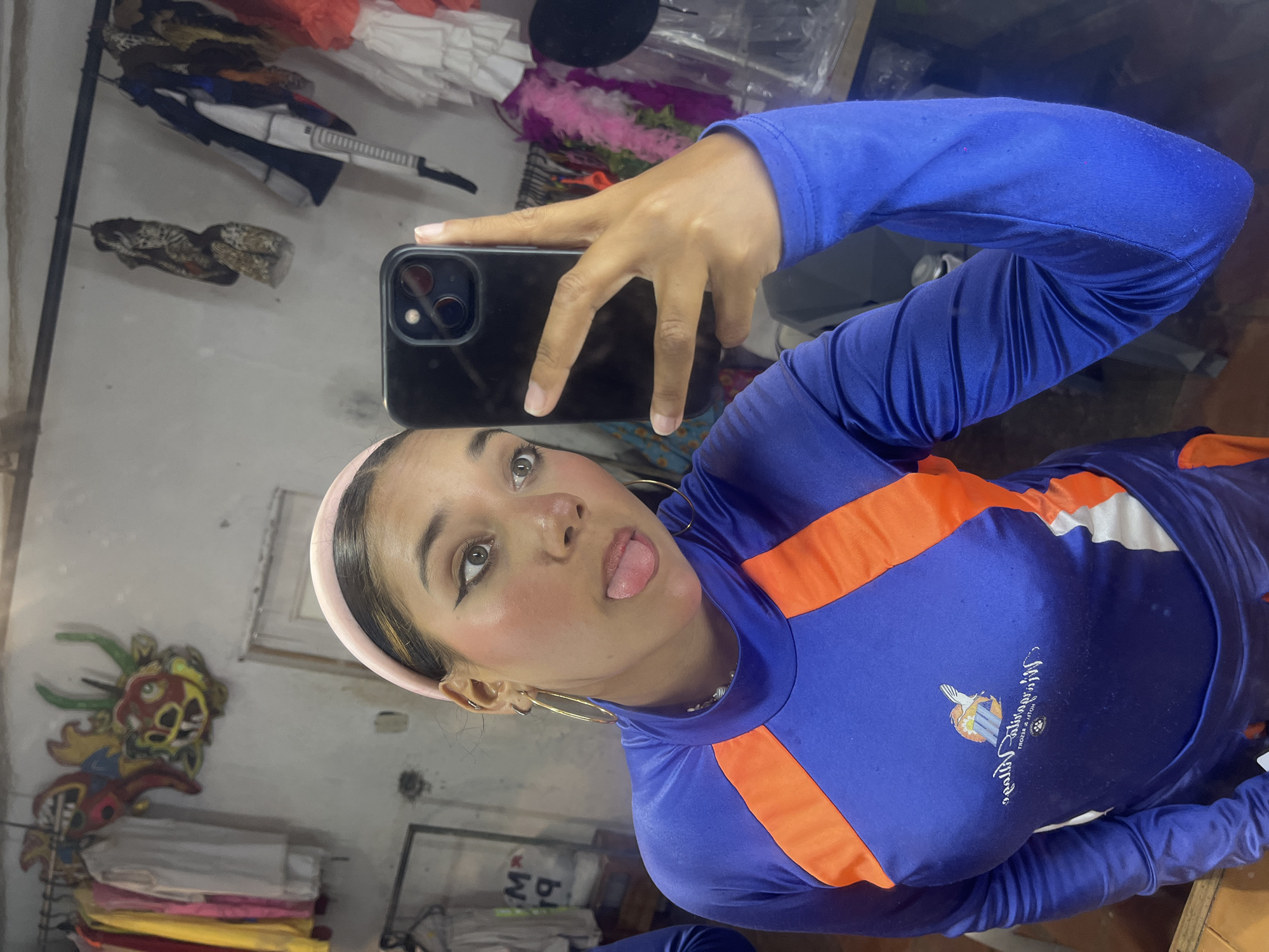 A young woman takes a mirror selfie indoors, showing a playful expression with her tongue sticking out. She has light skin, dark hair styled back with a pink headband, and is wearing makeup with prominent eyeliner and blush. Large golden hoop earrings adorn her ears. She holds a black smartphone, likely an iPhone, with her left hand to capture the reflection.
She is dressed in a vibrant royal blue, long-sleeved athletic or swim shirt, featuring distinctive orange panels across the chest and arms. On the front of her shirt, there's a mirrored logo. The logo appears to depict a bird, some buildings or structures, and text that seems to read "Salinas del Rey" or similar, followed by "Beach & Resort" or "Club & Resort," along with a small paw print symbol.
The background, visible in the mirror's reflection, suggests an eclectic indoor setting, possibly a market stall, shop, or a cluttered room in Porlamar, Venezuela. Various items are visible: rolls or stacks of colorful fabrics (white, red, pink, yellow), a pink feathery boa, and what appears to be a decorative, brightly colored dragon or monster mask in red, green, and yellow mounted on a wall. Another indistinct object resembling a weapon or tool hangs further back. On a dark blue surface in the lower background, mirrored white text is partially visible, possibly "M-BUI". The overall mood is casual and expressive.