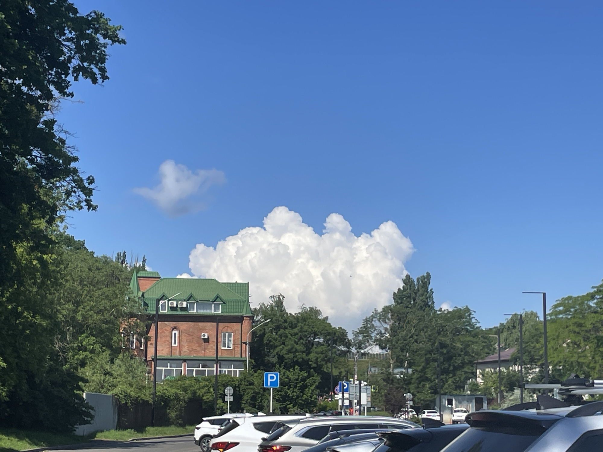The image depicts a street scene in Stavropol, Russia, on a sunny day with a bright blue sky and scattered white clouds. In the foreground, several cars are parked, with the closest ones being silver and white SUVs. A red brick building with a green roof is visible to the left, featuring multiple windows and air conditioning units. Adjacent to the building is a lush green area with trees and bushes. Two blue parking signs are also present. The background shows more trees and buildings, and the overall atmosphere suggests a typical urban environment.