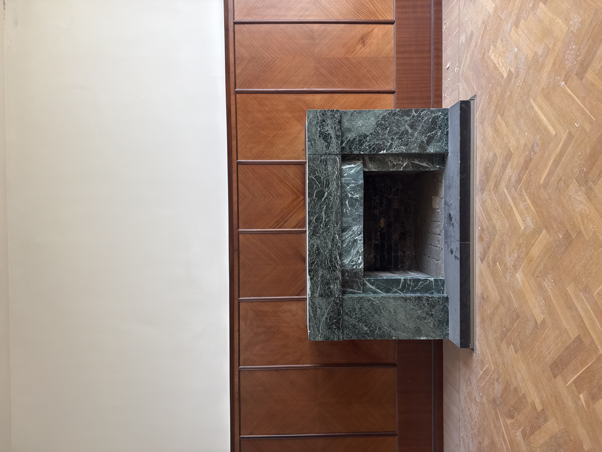 An indoor shot taken in Moscow, Russia, showing a room interior with a fireplace. The fireplace has a green marble surround. It is adjacent to a wooden wall and a light-colored wall. The floor is made of wood arranged in a herringbone pattern. There are no people present in the image.