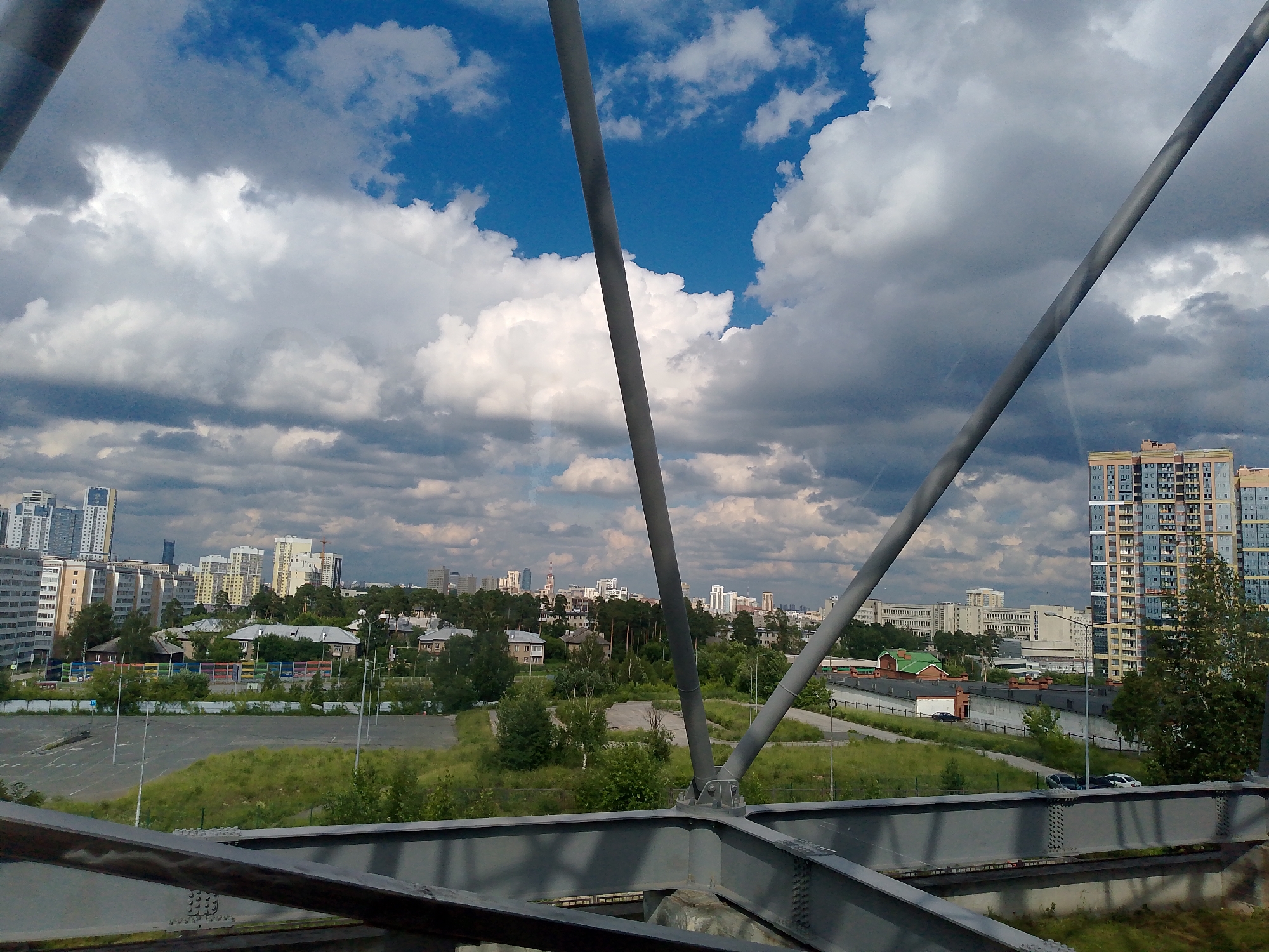The image captures a view of a city skyline under a partly cloudy sky, viewed from a metal structure. In the foreground, a grey metal beam with rivets is visible, suggesting the perspective is from a bridge or elevated walkway. The midground reveals a landscape with a large, paved, empty parking lot on the left, bordered by green grass and trees. Further into the midground, there are low-rise buildings with pitched roofs, and a long, colorful fence. A tall, modern apartment building with many windows and a blue facade is prominent on the right side of the image, with a green-roofed building and a stone wall in front of it. The background showcases a dense urban area with numerous tall buildings of varying architectural styles and heights, interspersed with trees and greenery. The sky is a mix of blue patches and large, dramatic grey and white clouds. Two large grey metal support beams, angled and converging towards the top of the frame, divide the scene, hinting at the structure from which the photograph was taken. A few cars are visible on the roads within the city.