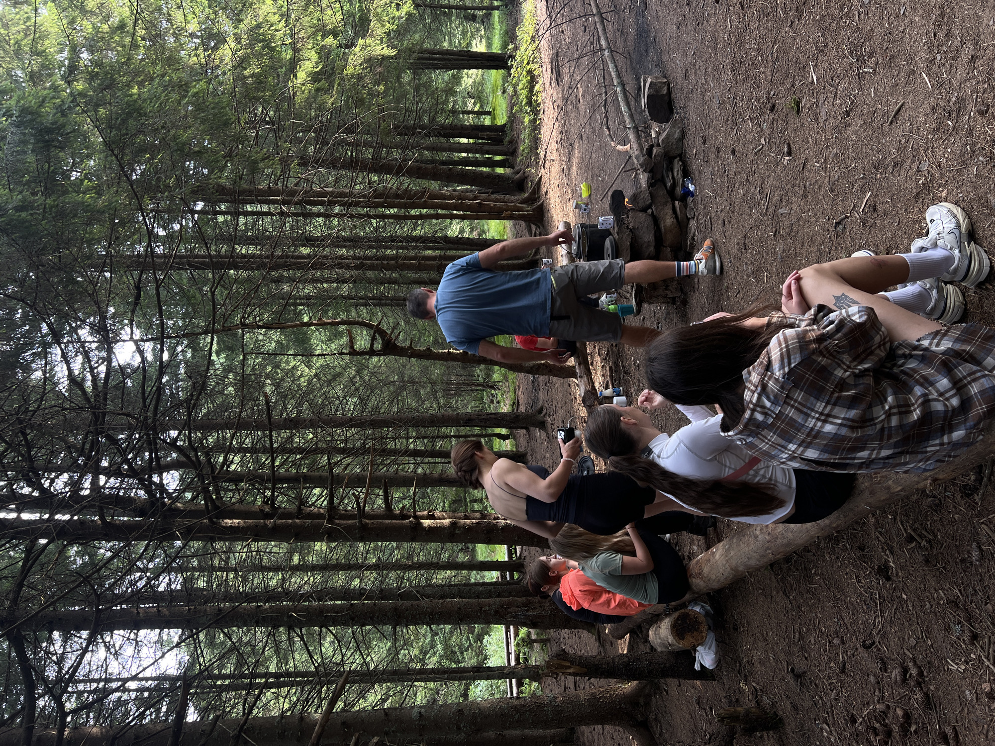 In a dense forest with tall pine trees, a group of people are gathered around a campfire. Several individuals are seated on a fallen log in the foreground, facing away from the camera. A man in a blue t-shirt and shorts stands near the campfire, which is built with rocks. Various cooking pots and containers are placed around the fire. The ground is covered with pine needles and dirt. In the background, a wooden structure, possibly a bridge or walkway, can be seen through the trees. The scene suggests an outdoor camping or gathering activity in a natural, wooded environment. The location is identified as the city of Vorokhta, Ukraine.