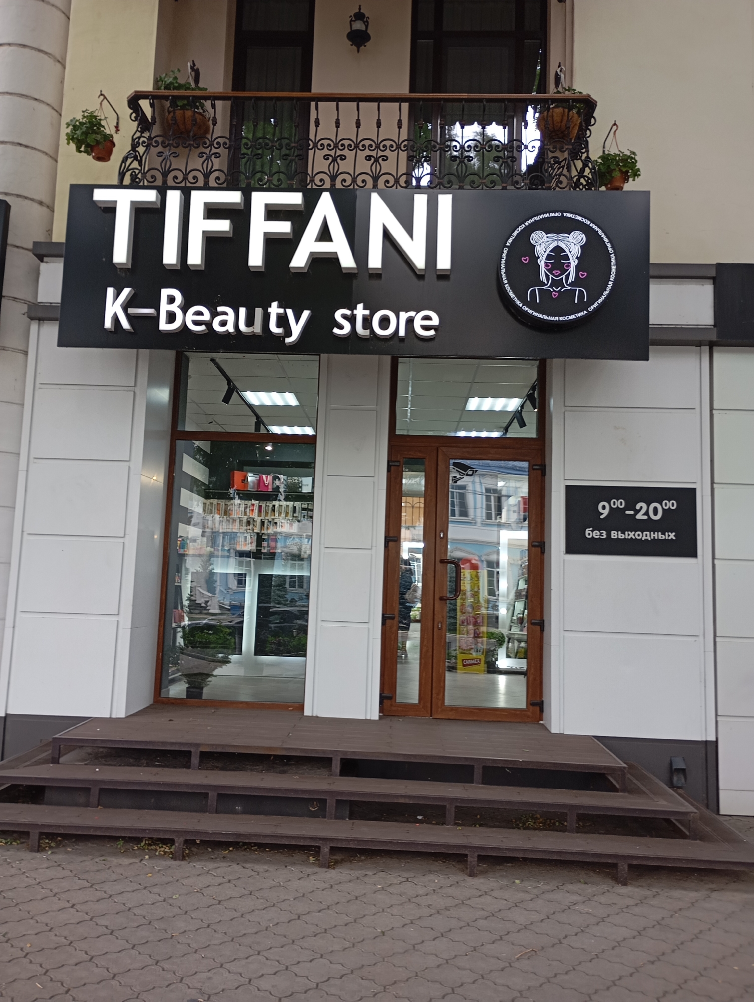 The image shows the storefront of a beauty store named "TIFFANI K-Beauty store" located in Tiraspol, Moldova. The store has a large black sign with white lettering on the facade, and a circular logo with a drawing of a woman's head and hearts. The entrance to the store features double glass doors, and a ramp leads up to the entrance. To the right of the entrance, there is a black sign indicating the store's operating hours as 9:00-20:00, without days off. The surrounding area appears to be a sidewalk paved with interlocking concrete blocks. There are plants on a balcony above the store.