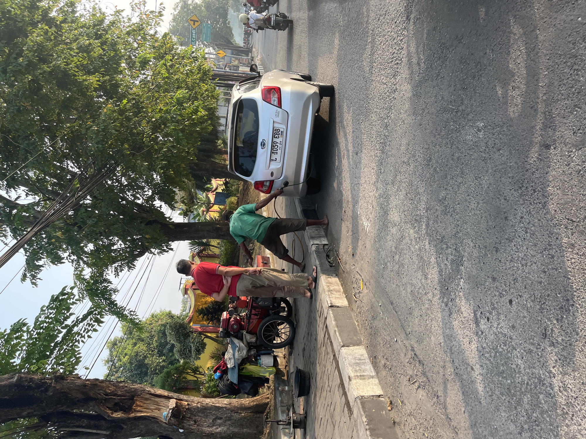 In Tenggulunan, Indonesia, two men are engaged in a roadside service. One man, wearing a red shirt and beige pants, stands with his back to the camera near a cart loaded with what appears to be tire repair equipment, including a red engine and various tools. The second man, in a green shirt and brown shorts, is actively attending to the rear of a silver Datsun car, identified by its license plate "L 1059 EBB". He is holding a hose that is connected to the car's fuel intake. The scene unfolds on a paved road adjacent to a sidewalk with a concrete curb, lined with trees and utility poles. The sun is casting long shadows, indicating it is either early morning or late afternoon. In the background, traffic can be seen, including motorcycles. The overall impression is one of routine urban activity, with a focus on automotive maintenance or assistance.