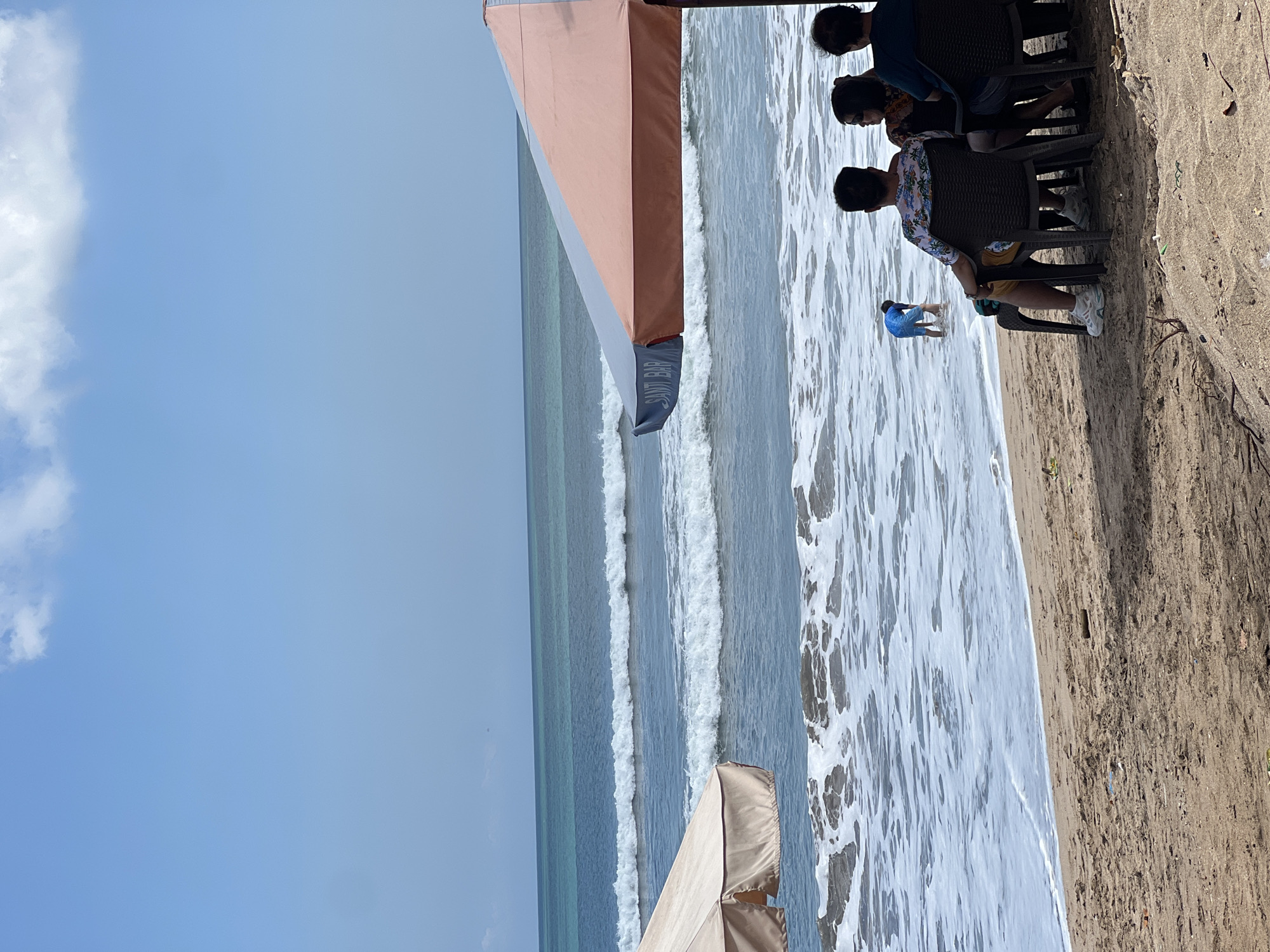 In Kuta, Indonesia, a beach scene unfolds under a clear blue sky with sparse clouds. The turquoise ocean displays multiple rows of white-capped waves, indicating a moderate swell. On the sandy shore, several people are seated on plastic chairs, facing the water. In the foreground, the backs of four individuals are visible, two men and two women. One man is wearing a floral shirt and yellow shorts, while another wears a dark blue shirt. To their right, a woman in a patterned top and another in a dark shirt are seated.
Further down the beach, a lone figure, a young person wearing a blue shirt and shorts, stands in the shallow water. Two large beach umbrellas provide shade; one is partially visible on the left, and another, a two-toned umbrella with orange and gray sections, is prominent on the right. The umbrella on the right has the text "SANTU BAR" printed on its side. The sand is a light brown, with some scattered debris. The overall atmosphere is one of relaxation and enjoyment of a beachside setting.