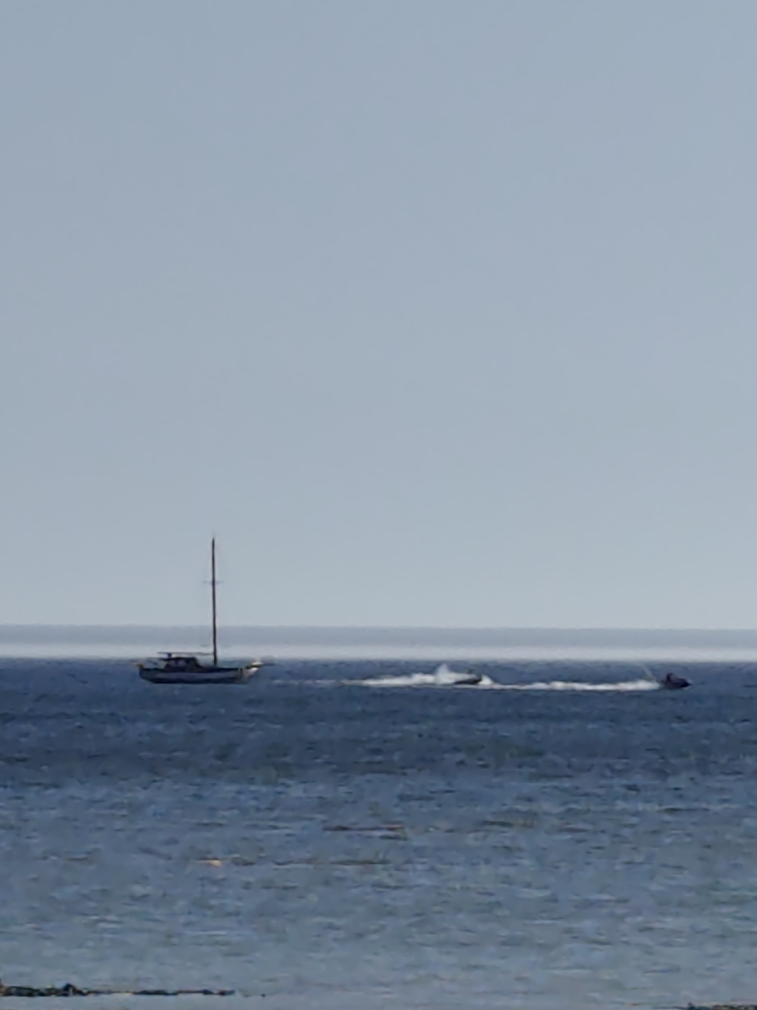 A sailboat with a tall mast is positioned in the middle of the frame, sailing on calm, deep blue water under a clear, pale blue sky. To the right of the sailboat, two jet skis are visible in the distance, creating white wakes as they move across the water. The horizon line is faint and appears to be very distant. The overall scene suggests a peaceful day at sea. This image depicts a view of the Caspian Sea near Aktau, Kazakhstan, an area known for its coastal scenery and recreational water activities.