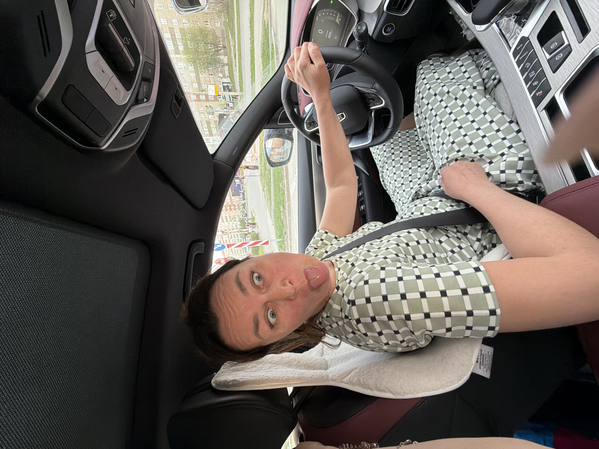 A middle-aged woman is in the driver's seat of a car, making a face and sticking her tongue out. She is wearing a patterned shirt and a seatbelt. The car is on a city street in Izhevsk, Russia, with buildings visible in the background. The interior of the car is visible, including the steering wheel, dashboard, and center console. A white pillow is behind the woman.