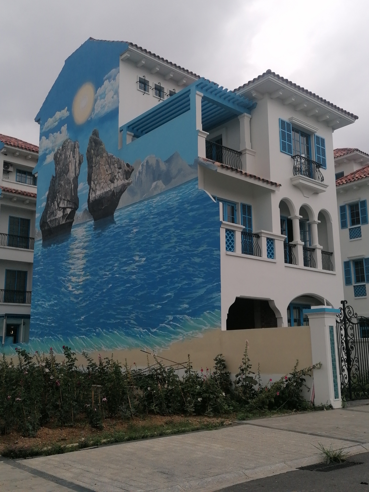The image presents a striking view of a multi-story building featuring a large, vibrant mural, set under an overcast sky in Bai Chay Ward, Vietnam.
The building itself is predominantly white, showcasing a Mediterranean or Moorish architectural style. It features a red tiled roof, white stucco walls, multiple windows adorned with bright blue louvered shutters, and black wrought-iron balconies. Decorative elements include arched windows and doorways, pillars, and a light blue wooden pergola structure extending from the upper levels. On the far left, a section of a similar building is partially visible.
The most prominent feature is the expansive mural painted on the left-facing wall of the building. It depicts a picturesque seascape: a clear blue sky with white clouds and a brilliant yellow sun. Two dramatic, rugged rock formations, strongly resembling the karst landscapes of Ha Long Bay, emerge from deep blue water. The water surface below them shows a shimmering reflection of the sun and distinct waves. A narrow strip of sandy beach with gentle blue and white waves is depicted at the bottom of the mural.
In the foreground, a verdant strip of greenery, composed of bushes dotted with light pink and white flowers, borders the building's base. Below the vegetation, a light gray concrete sidewalk with a gridded pattern leads towards a darker paved road. On the right, a white pillar with blue tile accents and an ornate black wrought-iron gate mark the entrance to the property.
The actual weather appears to be overcast, casting a soft, diffused light on the scene, which contrasts with the bright, sunny day portrayed in the mural. There are no people visible, nor is there any discernible text in the image. The architectural style and the specific geological features depicted in the mural are consistent with the coastal resort environment of Bai Chay Ward, Vietnam.