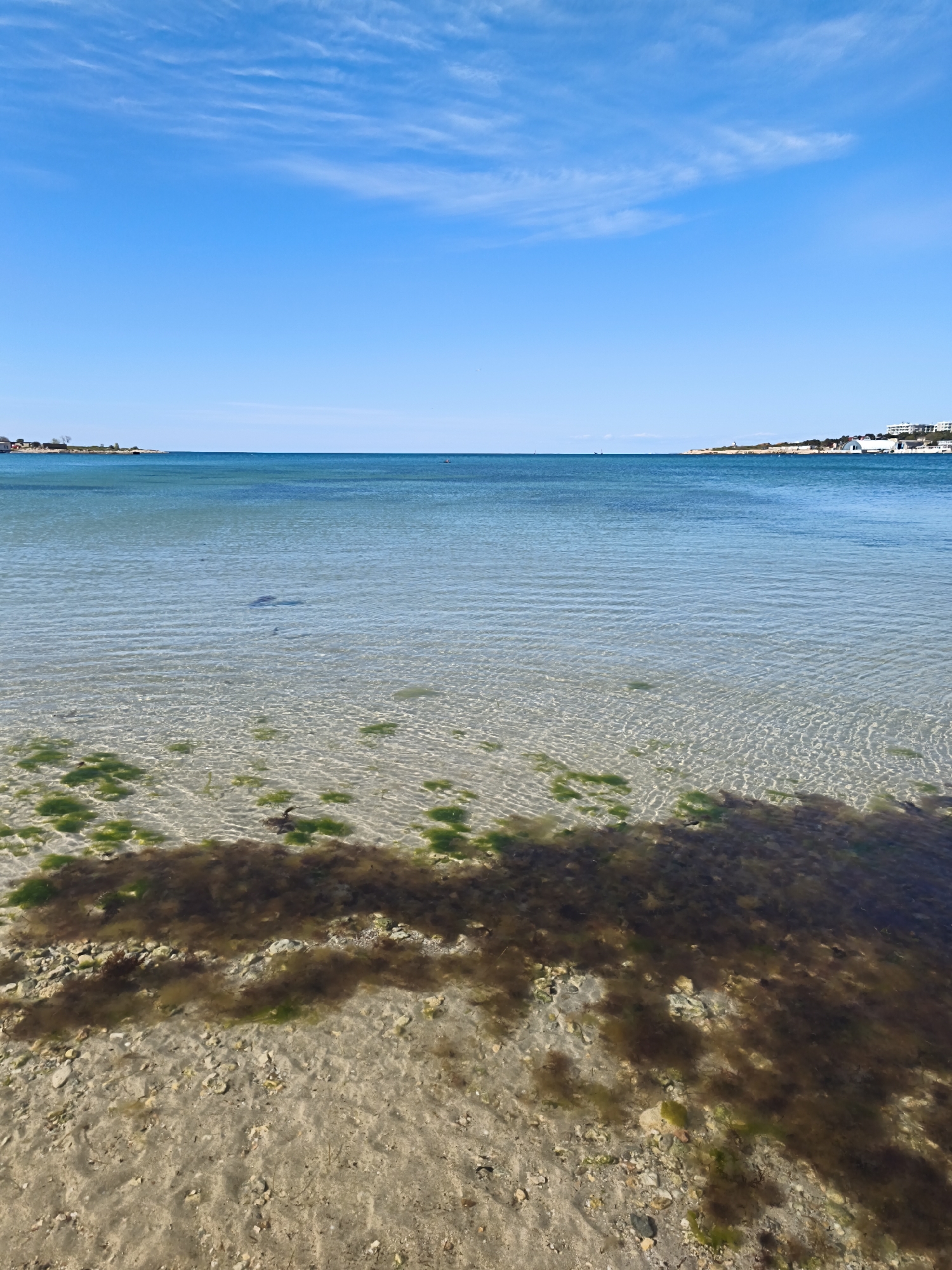 This media file presents a tranquil coastal landscape under a brilliant, clear blue sky dotted with a few wispy white clouds. The scene is dominated by a body of water, likely a sea or a large bay, with remarkably clear, calm water in the foreground. Through the transparent water, the seabed is distinctly visible, composed of light-colored sand and scattered small rocks. Patches of vibrant green and dark brown seaweed or algae grow on the bottom, adding natural texture to the aquatic foreground. Subtle ripples on the water's surface reflect the bright sunlight.
As the water extends further from the viewer, it gradually deepens in color to a richer blue. A faint, dark shape is discernible beneath the surface in the mid-ground, possibly a rock formation or a larger growth of submerged vegetation. The water stretches out to a distant horizon.
On the left side of the frame, a distant landmass features a few small buildings and some sparse trees, suggesting a residential area. To the right, another section of coastline is visible, showing more significant development with several prominent white buildings, possibly hotels or resorts, and large, curved white structures that appear architectural or industrial. These distant shores frame the expanse of water, characteristic of coastal areas such as Sevastopol, Ukraine.
The image conveys a sense of serenity and natural beauty, captured on a sunny day with pleasant weather. No people or specific activities are visible, emphasizing the peacefulness of the natural environment. The time of day is daytime, likely late morning or early afternoon, given the bright illumination. There is no visible text in the image.