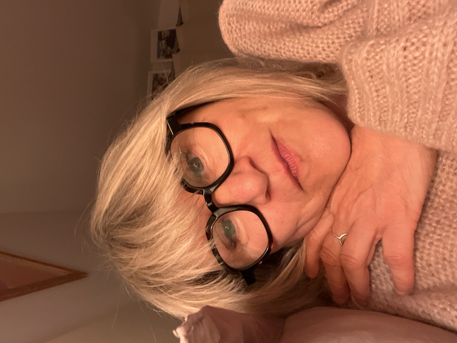 A woman with blonde hair and black-rimmed glasses peers at the camera. She's wearing a light pink sweater and has a ring on her finger. Her hand rests against her cheek. The image is softly lit, creating a warm tone. A partially visible picture frame is visible in the bottom left corner, and small photos hang on the wall behind her. The picture is taken at an angle, giving a slightly tilted perspective.