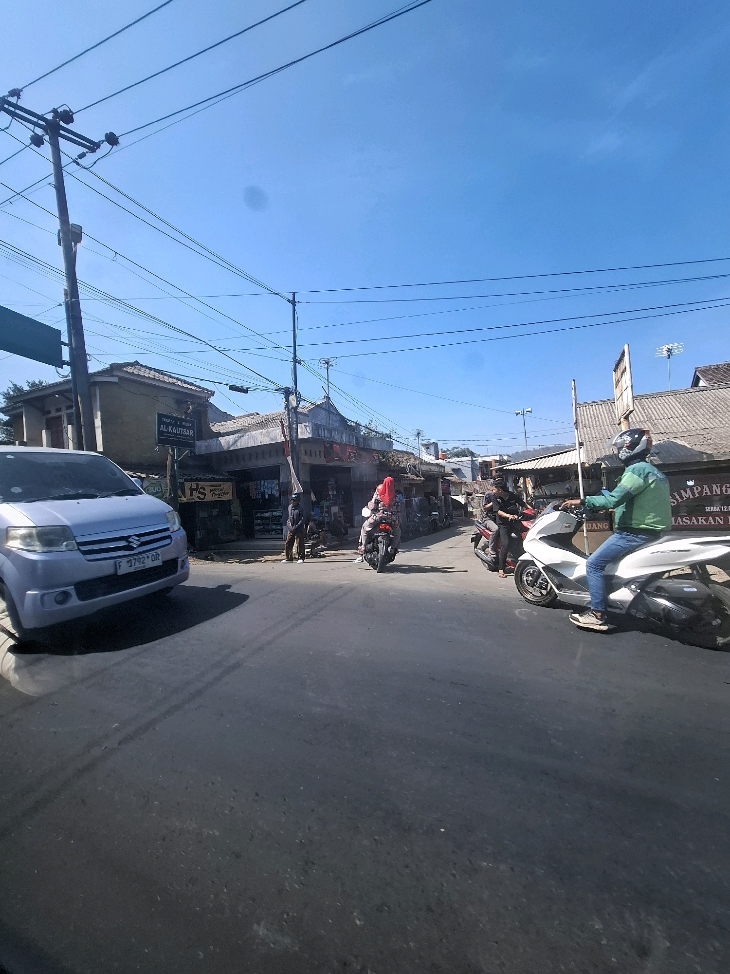 The image captures a street scene in Lembursawah, Indonesia, on a clear day with a bright blue sky. The road is paved and appears to be a two-lane street with visible tire marks. Several vehicles are present, including a light blue minivan on the left side of the frame, a white scooter in the right lane, and two motorcycles in the background, one red and one darker colored.
There are several people in the scene. A person wearing a green jacket and jeans, with a helmet, is riding the white scooter. In the background, a person in a red hijab is riding a motorcycle, and another person is seated behind them. Two men are standing near the shops on the right side of the street, one in a brown outfit and the other in dark clothing.
The environment features buildings lining the street, which appear to be shops or small businesses. One building has a sign that reads "YAYASAN & HUTMA AL-KAUTSAR". Overhead, a complex network of power lines is strung between utility poles, indicating an urban or semi-urban setting. The architecture of the buildings suggests a local Indonesian style, with tiled roofs. The overall atmosphere is one of everyday street activity.