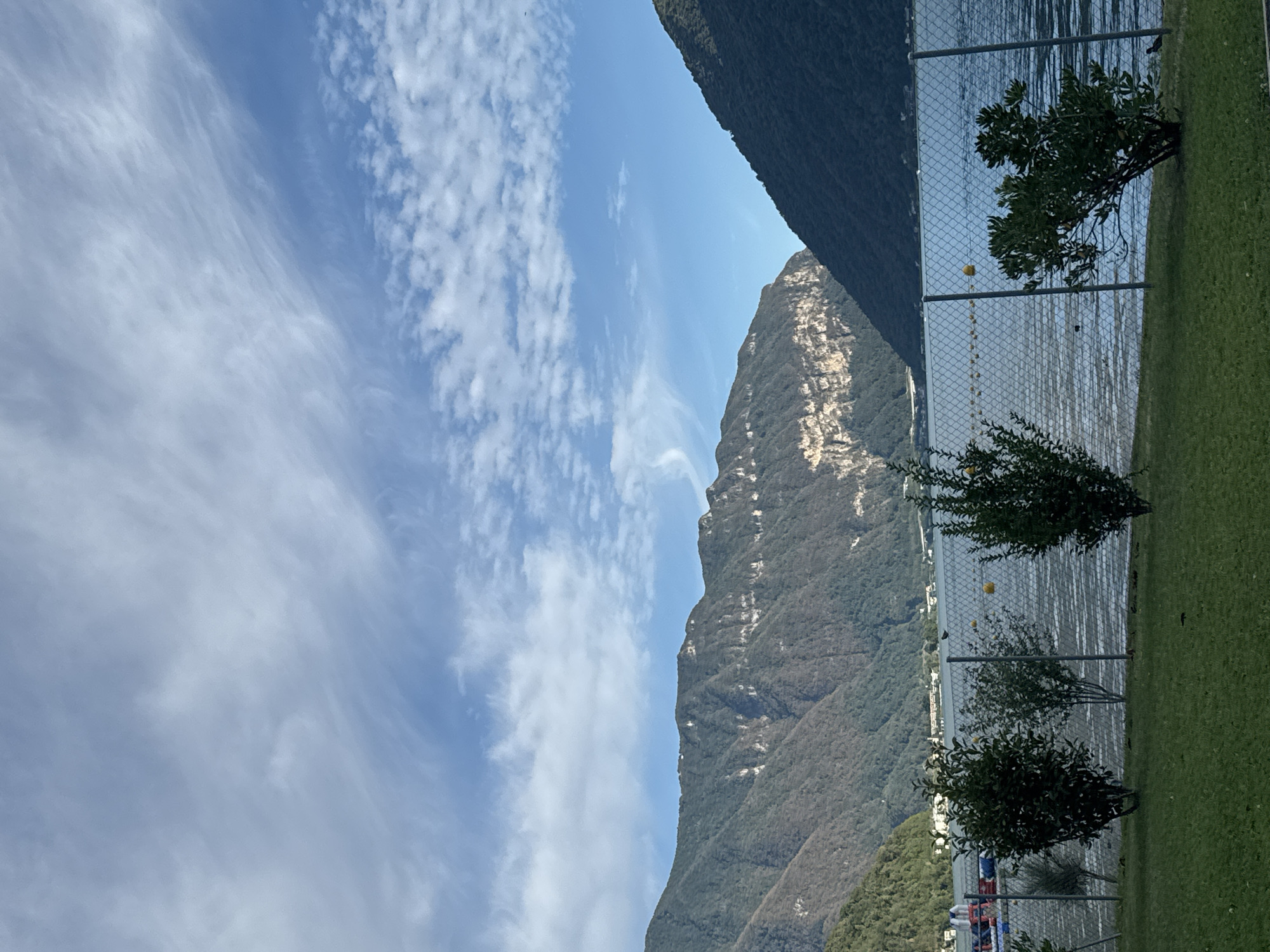 The image depicts a scenic view of Lake Lugano in Melide, Switzerland, with majestic mountains in the background under a partly cloudy sky. In the foreground, a well-maintained lawn is bordered by a chain-link fence, behind which several small trees with lush green foliage are planted. The lake itself is calm, with gentle ripples reflecting the sky and surrounding landscape. Bouys are visible floating on the water. In the distance, across the lake, the slopes of the mountains are covered with trees, and some buildings are nestled along the shoreline. The light suggests it is daytime, possibly late afternoon, given the angle of the sun and the shadows. The overall atmosphere is peaceful and serene, highlighting the natural beauty of the Swiss landscape.