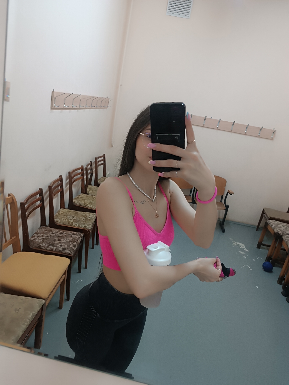 A young woman is captured taking a mirror selfie, visible from mid-thigh up. She has long dark hair and wears glasses, a bright pink ribbed sports bra or crop top, and black athletic leggings. Around her neck, she has a double-strand pearl necklace with a small, circular pendant. Her fingernails feature a pink and light blue gradient manicure, and she wears multiple rings. On her right wrist, she has a pink wristband. She holds a black smartphone, reflecting her face, in her right hand and a white water bottle in her left hand, along with a small pink object (possibly a hair tie) in her right.
The background, reflected in the mirror, suggests a changing room or a functional communal space within a gym or facility. Along the light-colored wall, there is a row of antique-looking wooden chairs with varied upholstered seats. Above the chairs, a wooden coat rack with several metal hooks is mounted. Another similar coat rack is visible further in the background on the right wall, along with what appears to be an air vent or grille high up. The floor is a light blue-grey color, with a noticeable spill of white powder or dust near a blue ball and other indistinct objects. The overall impression is that of a practical, unadorned interior. The time of day and weather are not discernible. No visible text is clearly legible.