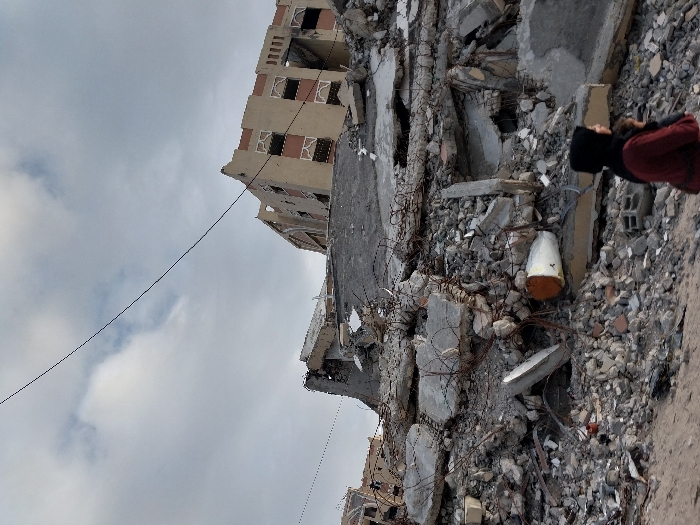 Amidst a scene of devastation, rubble and twisted metal cover the ground, indicative of a building collapse. In the background, a partially standing beige structure with windows is visible against a cloudy sky. A person with a dark head covering and a red shirt appears on the right edge of the frame. Debris including rebar and concrete fragments are scattered throughout. A conical object with a white top and orange bottom lies in the rubble.