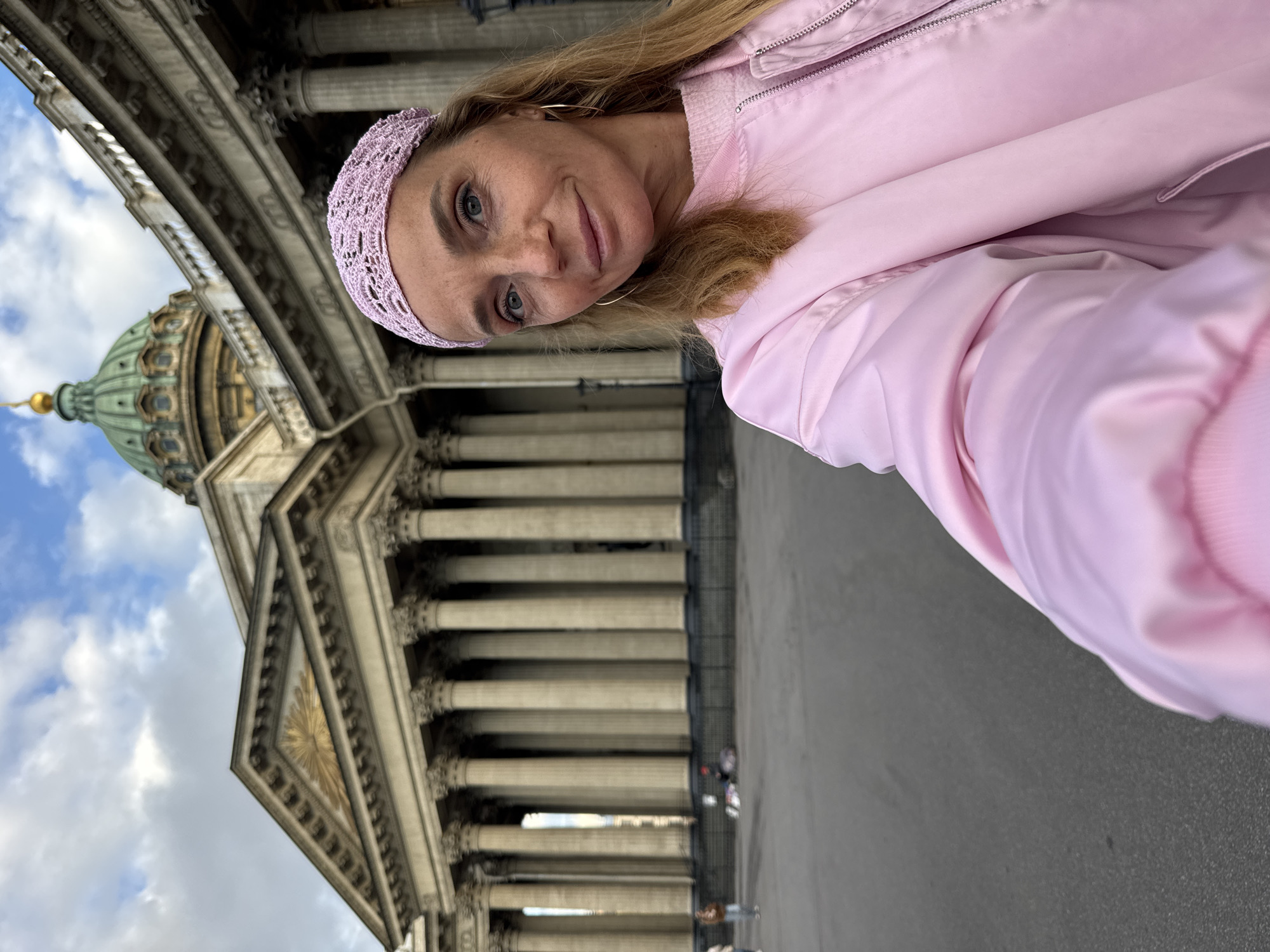 A woman with blonde hair and blue eyes is taking a selfie in front of a grand building. She is wearing a pink jacket and a pink crocheted headband. The building behind her has a large green dome with a golden spire, and a facade featuring rows of large columns and intricate architectural details. The sky above is blue with some white clouds. The ground in front of the building appears to be paved. Several other people are visible in the background, small in the distance, suggesting the scene is outdoors in a public space. The overall impression is one of sightseeing or enjoying a day out. The image is oriented sideways.