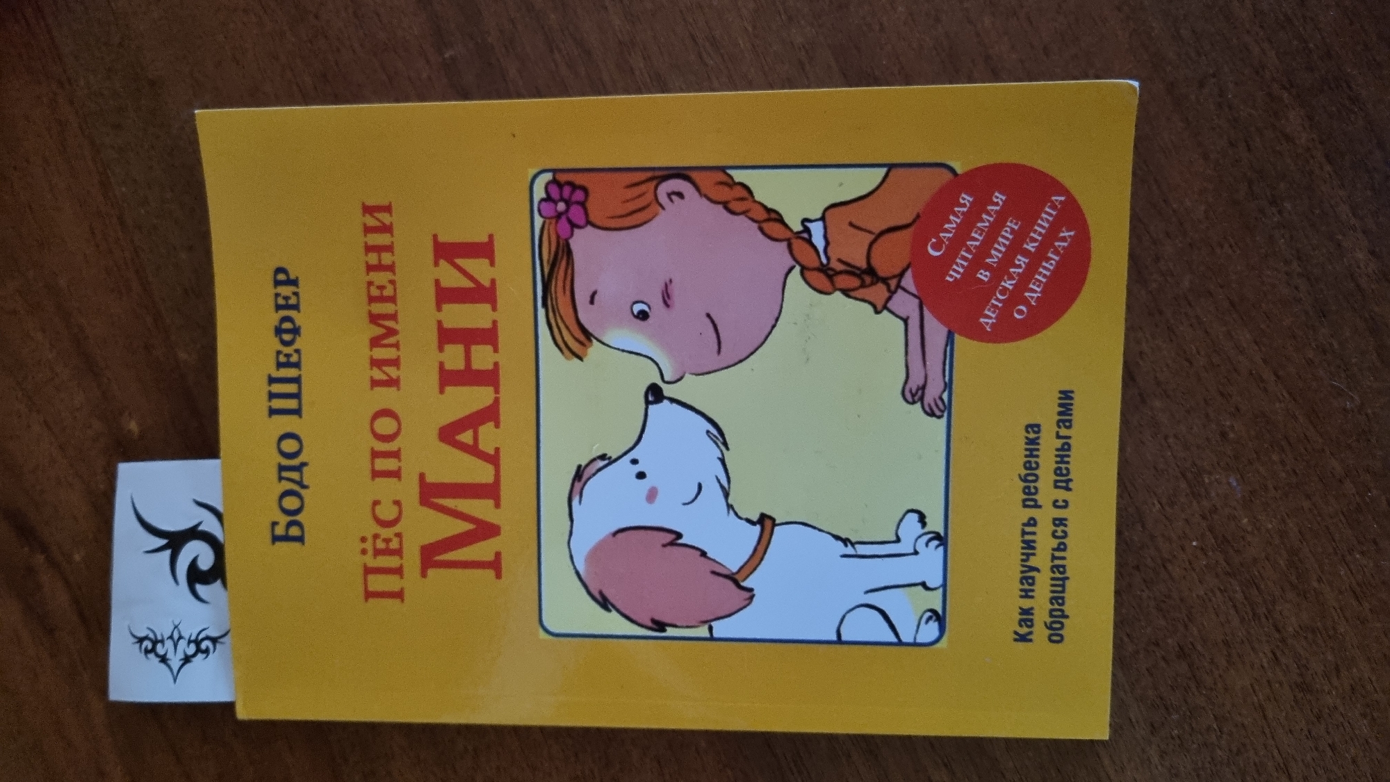 A close-up, overhead shot shows a bright yellow book titled "Пёс по имени Мани" (Dog named Mani) by Бодо Шефер (Bodo Schafer). The book is lying on a wooden surface, likely a table. A white bookmark with a black tribal tattoo design is peeking out from the top of the book. The book cover features an illustration of a happy cartoon dog looking at a girl with braided red hair, who is wearing an orange dress and a pink flower in her hair. Below the illustration, the text reads "Как научить ребенка обращаться с деньгами" (How to teach a child to handle money). A red circular sticker is placed on the lower right side of the book cover, with text that reads "САМАЯ ЧИТАЕМАЯ В МИРЕ ДЕТСКАЯ КНИГА О ДЕНЬГАХ" (The most read children's book about money in the world). The image does not provide any specific information or context related to the city of Солнечная Поляна-3 Снт, Russia.