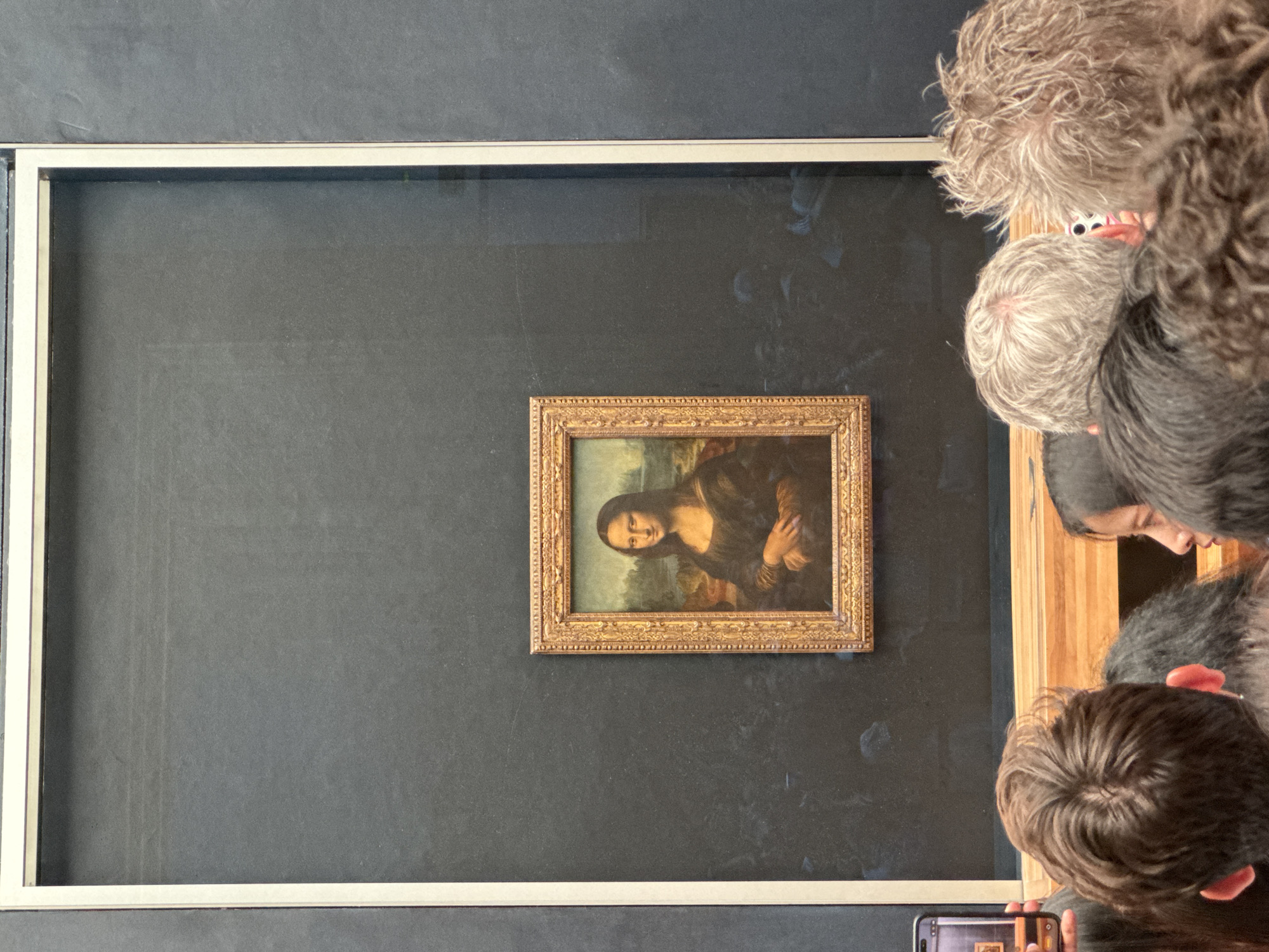 The image shows the Mona Lisa painting displayed in a museum. The painting is in a golden ornate frame and is behind a protective barrier. A crowd of people are gathered in front of the painting, many holding up their phones to take pictures. The museum appears to be the Louvre Museum in Paris, France. The painting is centered in the frame and takes up a significant portion of the image. The lighting is dim, with a spotlight focused on the painting. The crowd is dense, and the people are of various ages and backgrounds. The overall atmosphere is one of awe and admiration for the famous artwork.