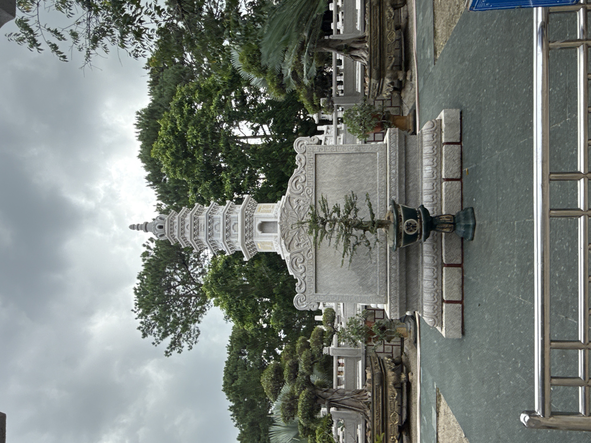 The image shows a serene outdoor setting, likely part of a temple or memorial complex in Da Lat, Vietnam. Dominating the center is a multi-tiered white pagoda, intricately carved, set against a backdrop of lush green trees and an overcast sky. In front of the pagoda is a substantial stone pedestal with decorative carvings. At the base of the pedestal sits a bonsai tree in a dark green ornate pot, a solitary green focal point. Flanking the pagoda, on either side, are large, ornately carved stone planters containing mature trees with gnarled trunks, suggesting they are ancient or carefully cultivated bonsai. The ground is covered in a dark green, textured mat, creating a uniform surface. Beyond the pagoda and planters, a stone balustrade lines the perimeter, with further greenery and hints of other structures visible in the background. A silver metal railing in the foreground frames the lower part of the scene, suggesting a visitor's viewpoint. The overall atmosphere is peaceful and contemplative.