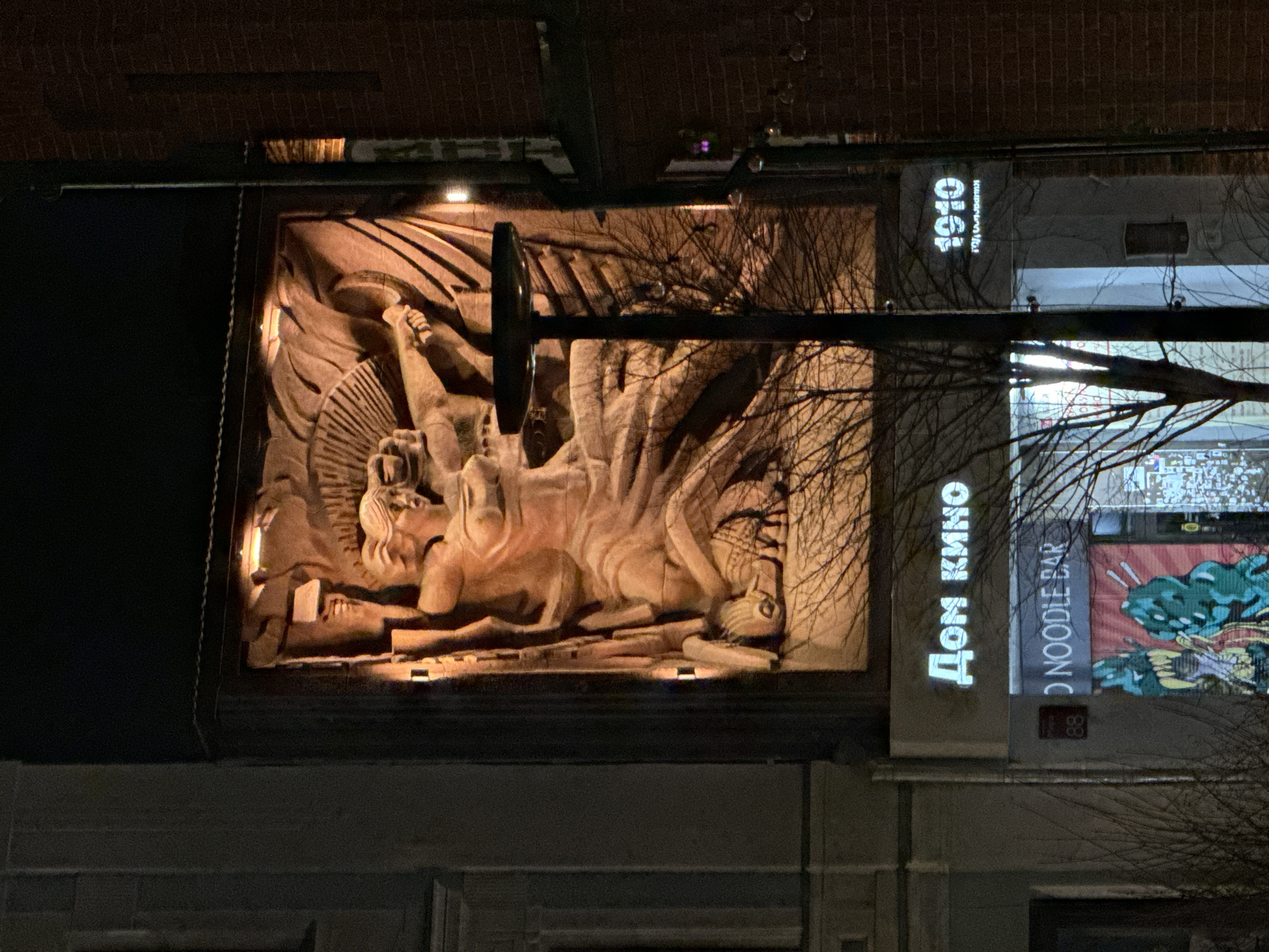 The image shows a building in Krasnoyarsk, Russia, at night. The main feature is a large, illuminated bas-relief carving depicting a mythological scene with human-like figures and an animal. To the right of the carving, a building has vertical signage with the words "ДОМ КИНО" (Dom Kino) and "NOODLE BAR" visible. The street address "88" is also visible. Adjacent to the Dom Kino is another storefront with the number "1910" displayed. A tree branch and a lamp post partially obstruct the view. The buildings are brick or stone. The lighting is artificial, illuminating the carving and the signs.