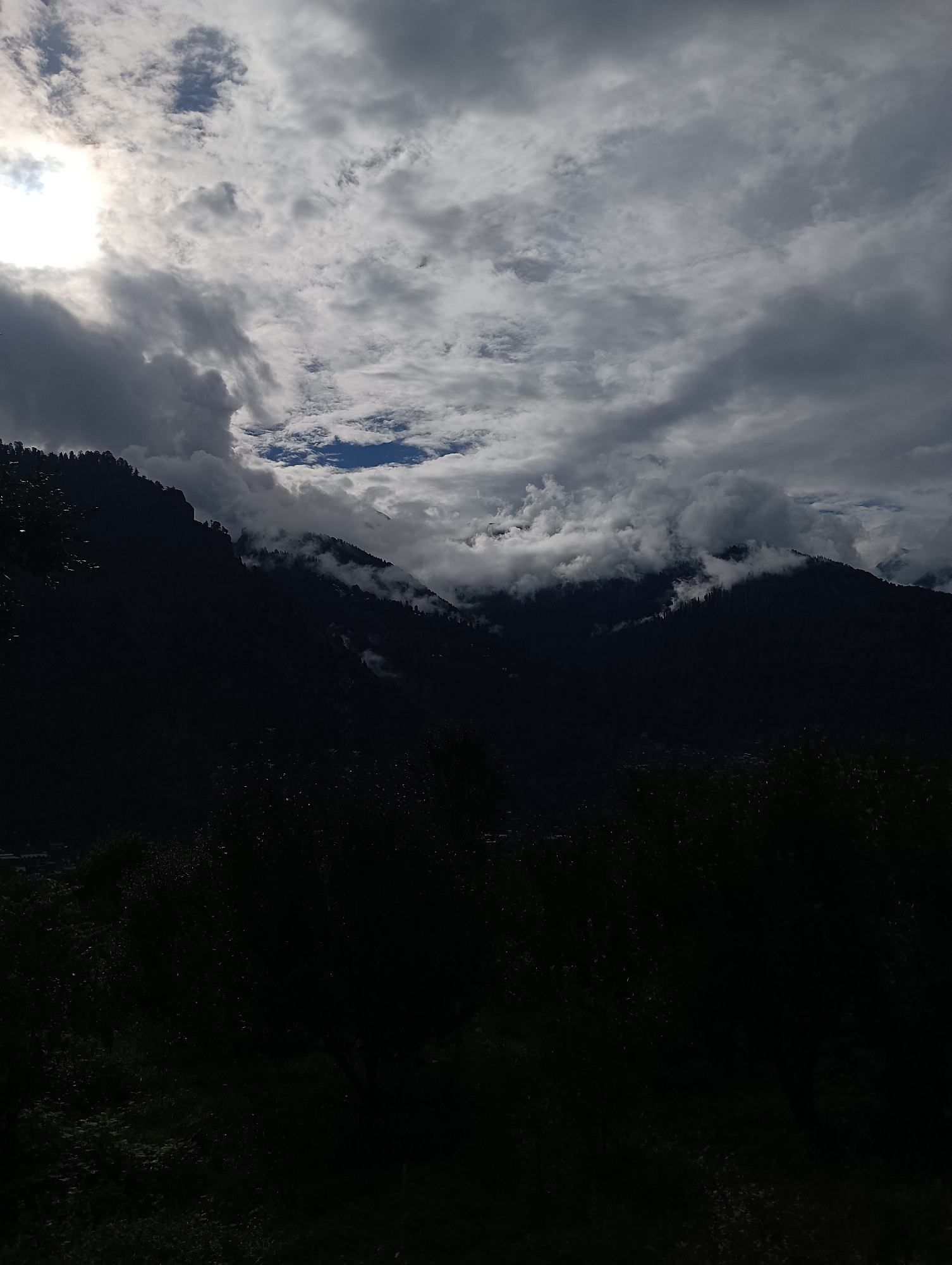 The image depicts a view of the city of Chhyal, India, characterized by its mountainous terrain and cloudy skies. The foreground is dominated by dark, silhouetted trees, obscuring much of the lower portion of the scene. Above the trees, the dark, imposing silhouettes of mountains rise steeply. These mountains are partially shrouded in dense clouds, which appear to be drifting and swirling around the peaks. The sky is a dramatic mix of dark grey storm clouds and patches of bright, diffused sunlight breaking through. In the upper left corner, a bright spot indicates the sun, partially obscured by clouds, casting a subtle glow on the scene. The overall impression is one of a moody and atmospheric landscape, with the natural beauty of the mountains and the dynamic weather conditions taking center stage. There are no visible people, specific structures, or activities to note.