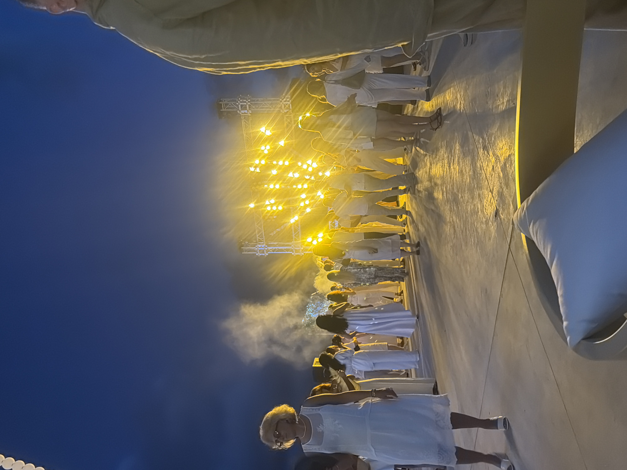An evening outdoor event is depicted in Shekvetili, Georgia. A crowd of people, predominantly dressed in white, are gathered in front of a large, brightly lit stage structure. The stage features multiple rows of yellow spotlights, creating a vibrant and energetic atmosphere. Smoke or fog emanates from behind the stage, adding to the dramatic effect. The ground is a light-colored, paved surface, reflecting the bright lights. In the foreground, on the left side of the image, a woman wearing glasses and a white dress stands looking to the left. On the right side of the image, a person in a light-colored jacket is partially visible. The sky above is a dark blue, indicating it is either dusk or night. The overall scene suggests a concert, festival, or similar public gathering.