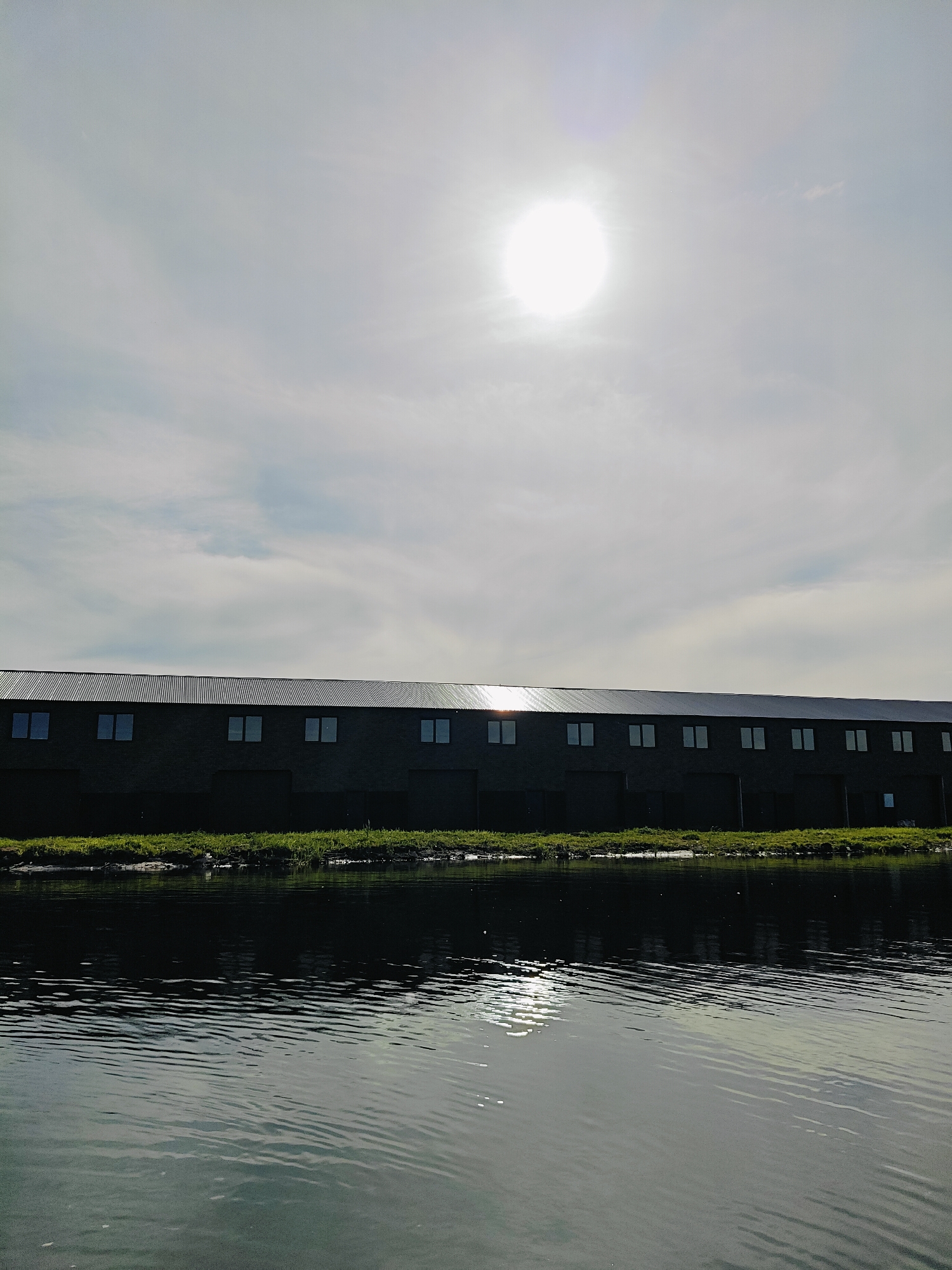 This image depicts a wide, calm body of water reflecting the sky and a long, dark building. The building appears to be a warehouse or industrial structure with a corrugated metal roof and a row of evenly spaced windows. A strip of green grass separates the building from the water's edge. The sun is visible in the sky, slightly to the left of the center, appearing as a bright, diffused orb through a hazy sky with scattered clouds. The overall impression is one of a serene, possibly industrial or agricultural landscape under a bright but overcast day. The water is smooth with gentle ripples, suggesting a lack of strong wind. The lighting indicates it is daytime, likely late morning or afternoon, with the sun high in the sky. There is no visible text in the image.