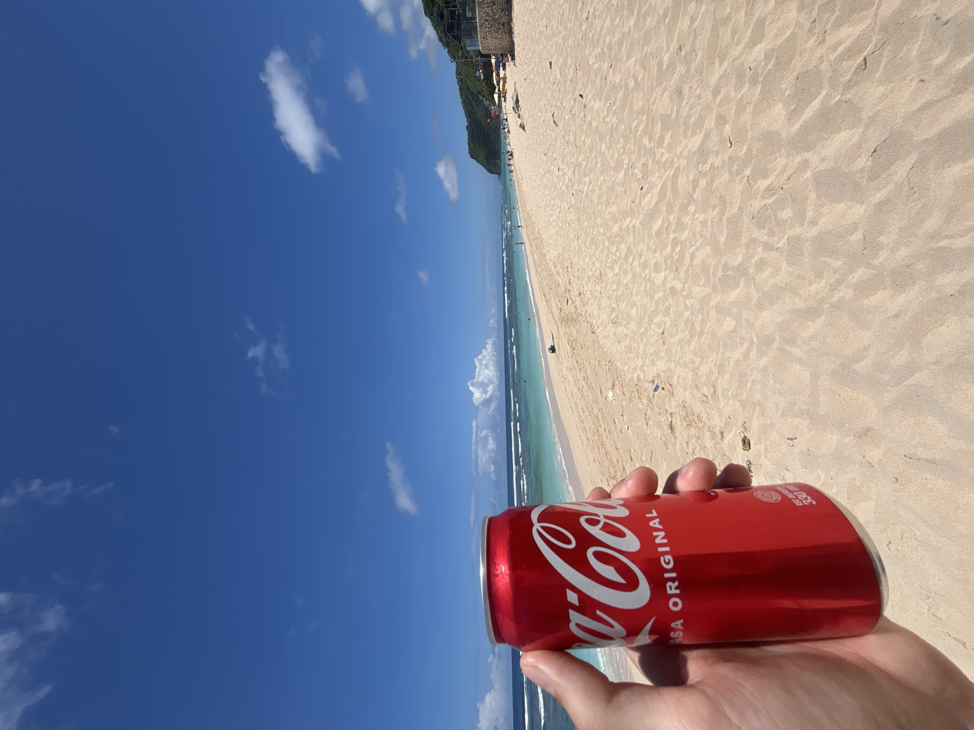 A Coca-Cola can is held by a person against the backdrop of a beach in Ungasan, Indonesia. The can, red with white lettering, reads "Coca-Cola" and "SA ORIGINAL". The person's fingers are visible holding the can. The beach has light-colored sand, and there are waves visible in the turquoise water. People can be seen in the distance on the beach. The sky is blue with scattered white clouds. A green, vegetation-covered cliff or hill is also visible in the background.