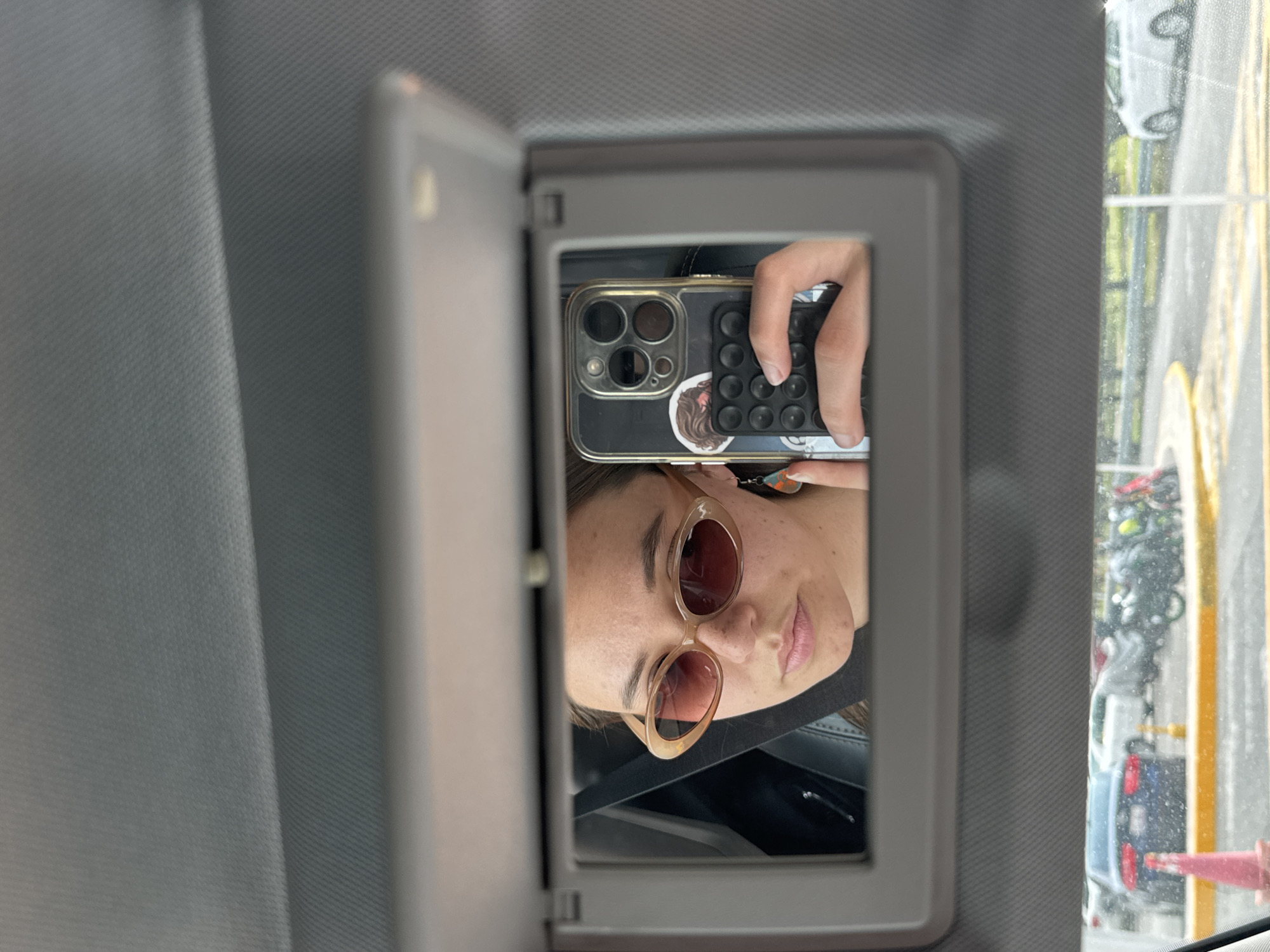 A woman takes a selfie in the mirror of a car visor. She wears sunglasses and holds a black iPhone with a pop-it case. The car is in San Andrés Cholula, Mexico, as indicated by the license plates and road markings visible through the window.