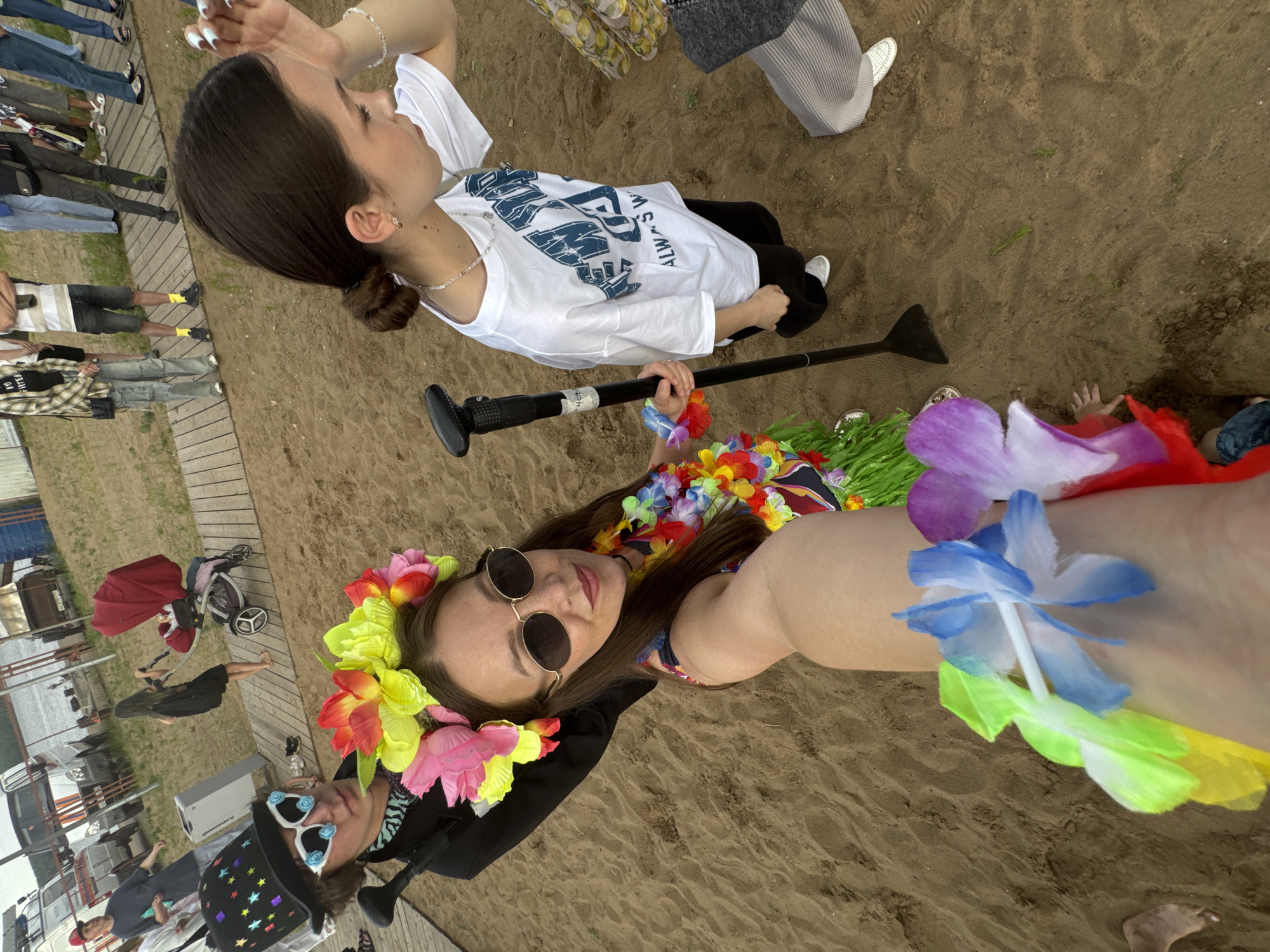 The image depicts an outdoor gathering on a sandy area, possibly a beach or park, located in the city of Chaikovsky, Russia. In the foreground, two individuals are prominently featured. One is wearing a black top hat adorned with colorful stars and novelty sunglasses, a black jacket, and a patterned scarf. This person is holding a black paddle or oar. Next to them, another individual wears a vibrant flower crown, sunglasses, and a leis, suggesting a festive or themed event.
Further in the background, several other individuals are visible. One person is pushing a red stroller with a baby. Other people are standing on a wooden boardwalk, some are casually dressed, while others appear to be in transit. In the far background, there are buildings or structures that might be associated with a recreational area or event venue, and a body of water is visible, potentially a river or lake. The overall scene suggests a social gathering with a mix of casual and themed attire, set against an urban backdrop with natural elements.