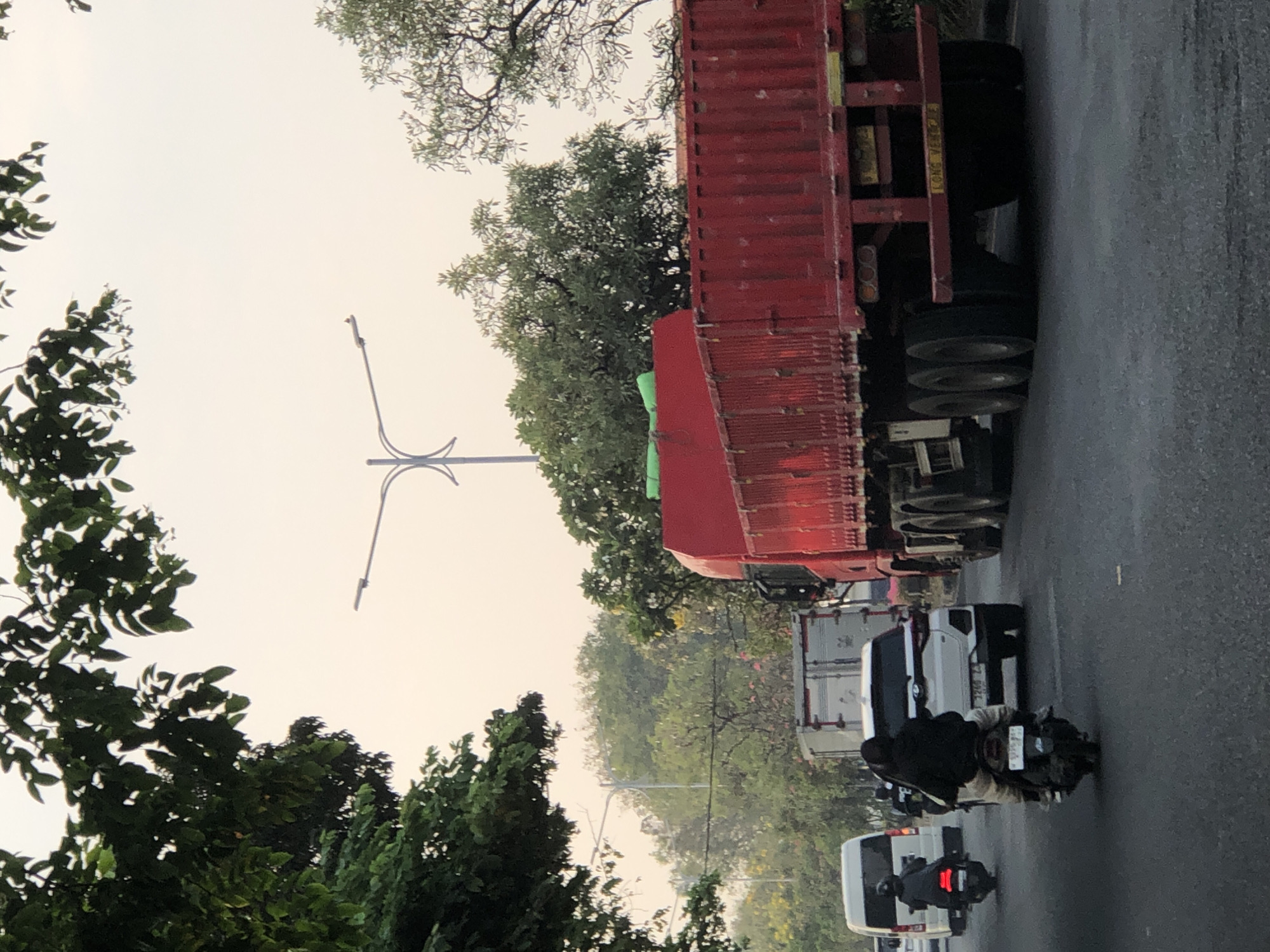 The image depicts a traffic scene on a road in Sumorame, Indonesia. In the foreground, a red flatbed truck with a green tarp over its cargo is prominently featured, occupying the right side of the frame. The truck has multiple axles and wheels. To the left of the truck, a white SUV is visible, followed by a motorcycle with two occupants. Further to the left, a white van is also present in the traffic. The road appears to be a multi-lane thoroughfare, with vehicles moving in the same direction. Trees line the sides of the road, with dense foliage in the upper left and scattered trees with some flowering branches further down the road. A street lamp with an unusual, almost abstract design stands in the background, above the traffic. The sky is overcast, suggesting a hazy or possibly early morning/late afternoon light. The overall impression is one of moderate traffic congestion on a paved road surrounded by greenery.
