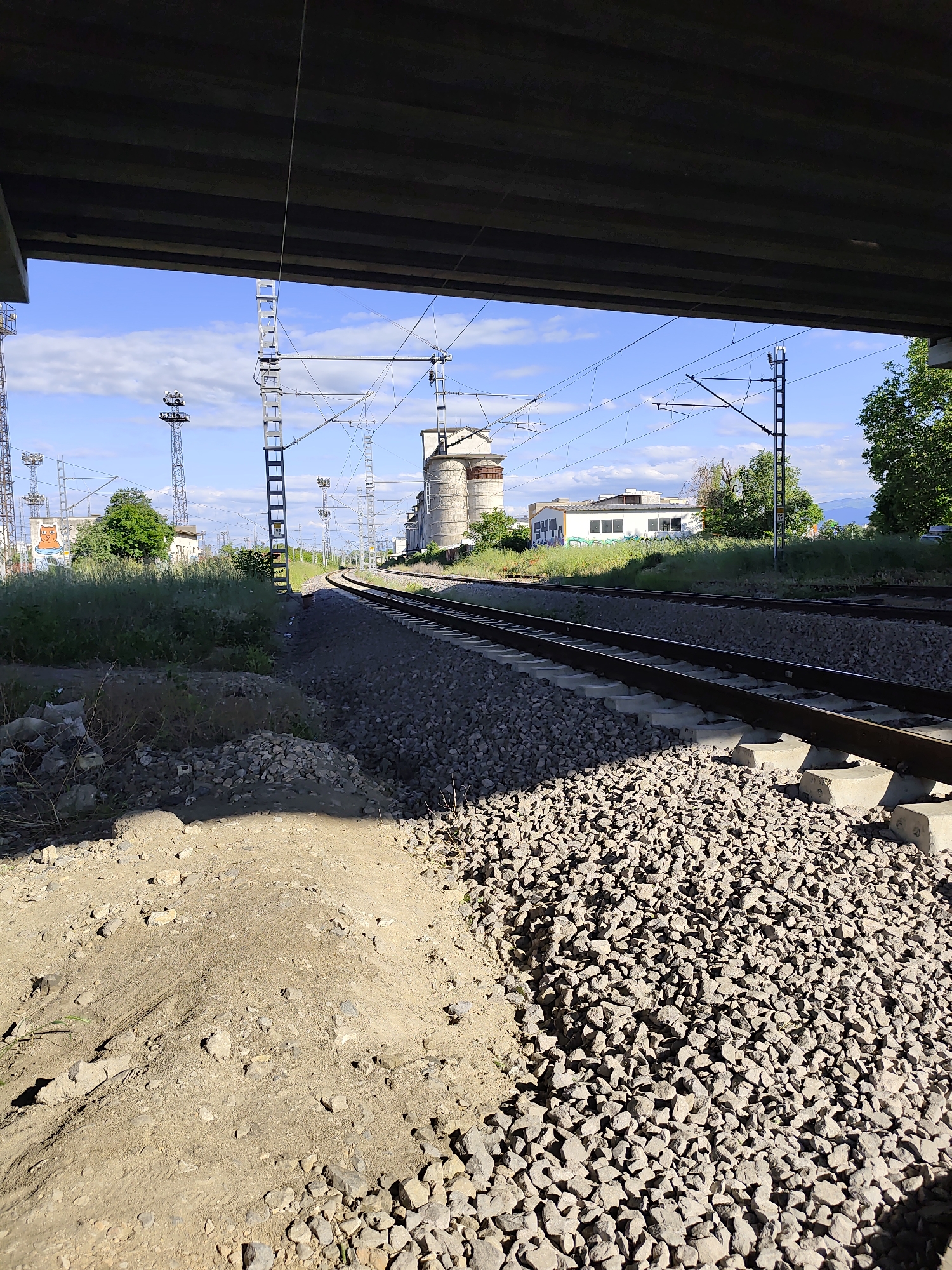 The image shows a section of railway tracks viewed from under a bridge in Plovdiv, Bulgaria. The tracks run roughly straight into the distance with overhead electrical wires and supporting structures present. A gray gravel ballast surrounds the tracks. To the left foreground is a sandy area. Beyond the tracks, there are industrial buildings and vegetation. The sky is blue with some scattered clouds. The image appears to have been taken during the day in natural light.